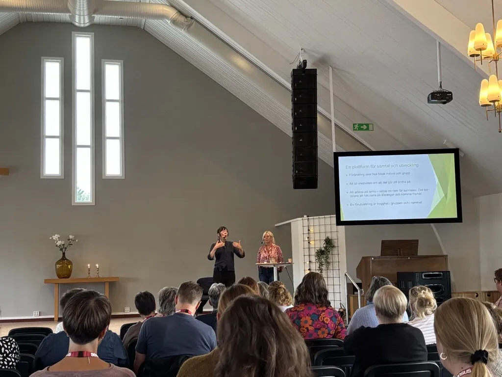 Två föreläsare håller en presentation framför en publik i en ljus lokal med högt i tak och vita väggar. En skärm visar en presentation med rubriken 'En plattform för samtal och utveckling'.