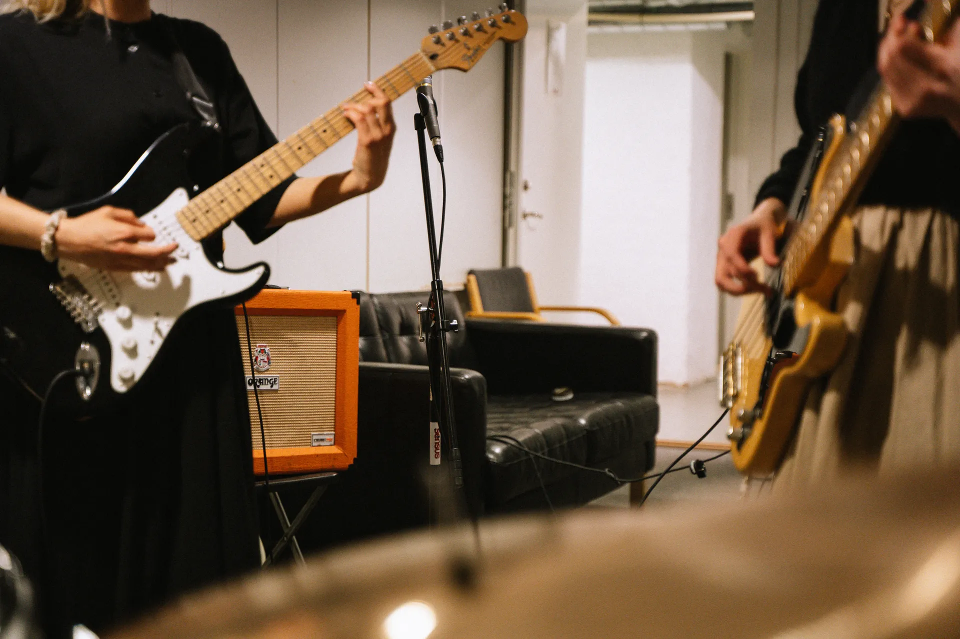 Två gitarrister spelar elgitarr i en replokal med en orange förstärkare, mikrofonstativ och en svart lädersoffa i bakgrunden