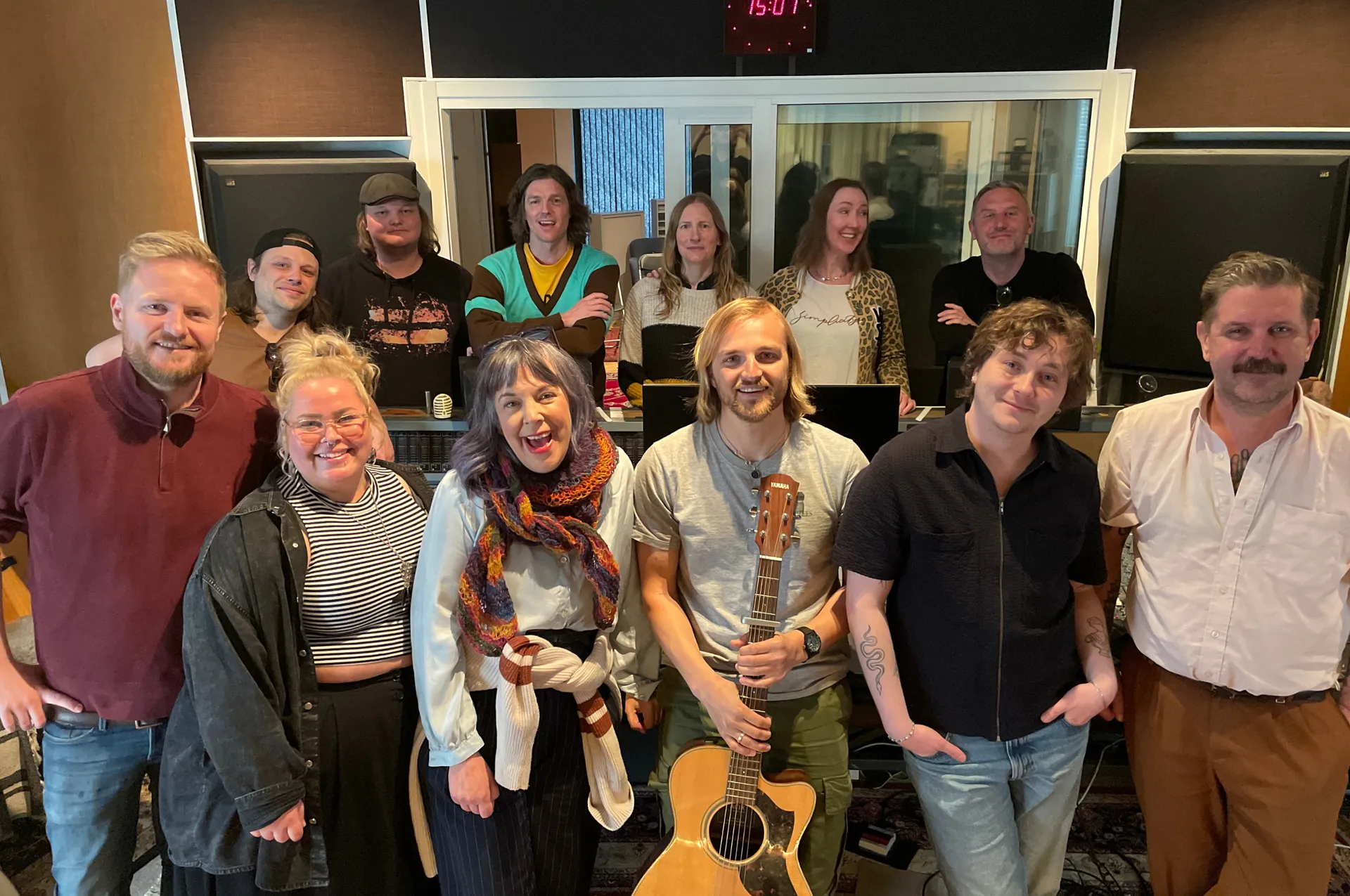 Gruppbild av tretton personer som poserar glatt i en inspelningsstudio, där en man i mitten håller en akustisk gitarr. I bakgrunden syns studioutrustning och ett kontrollrum bakom glasfönster.