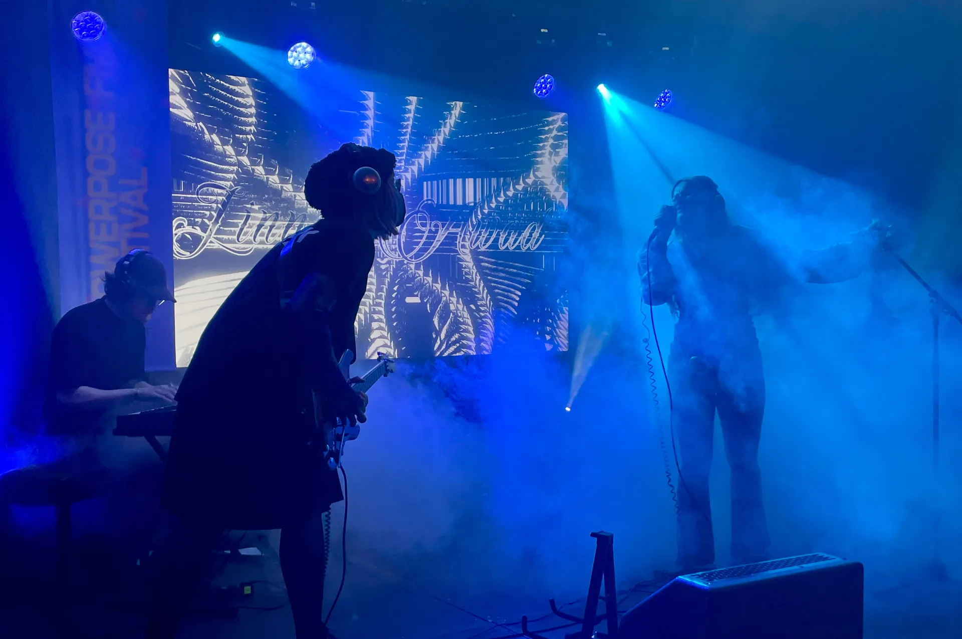 Konsertscen med blå scenbelysning och rökeffekter där musiker uppträder, en sångerska vid mikrofon och en gitarrist i silhuett mot en grafisk bakgrundsprojektion