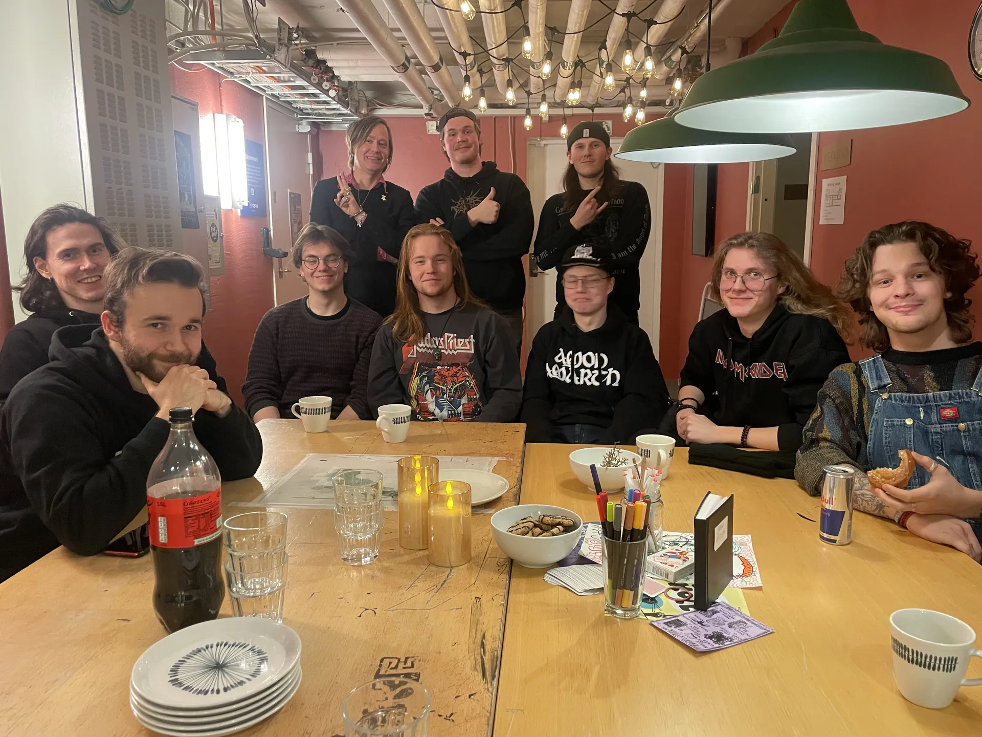 En grupp på elva unga personer samlade runt ett träbord i ett mysigt rum med röda väggar och industriellt tak. På bordet finns kaffekoppar, glas, ljus, en skål med kakor och pennor. Flera bär svarta tröjor med bandlogotyper och stämningen verkar avsl