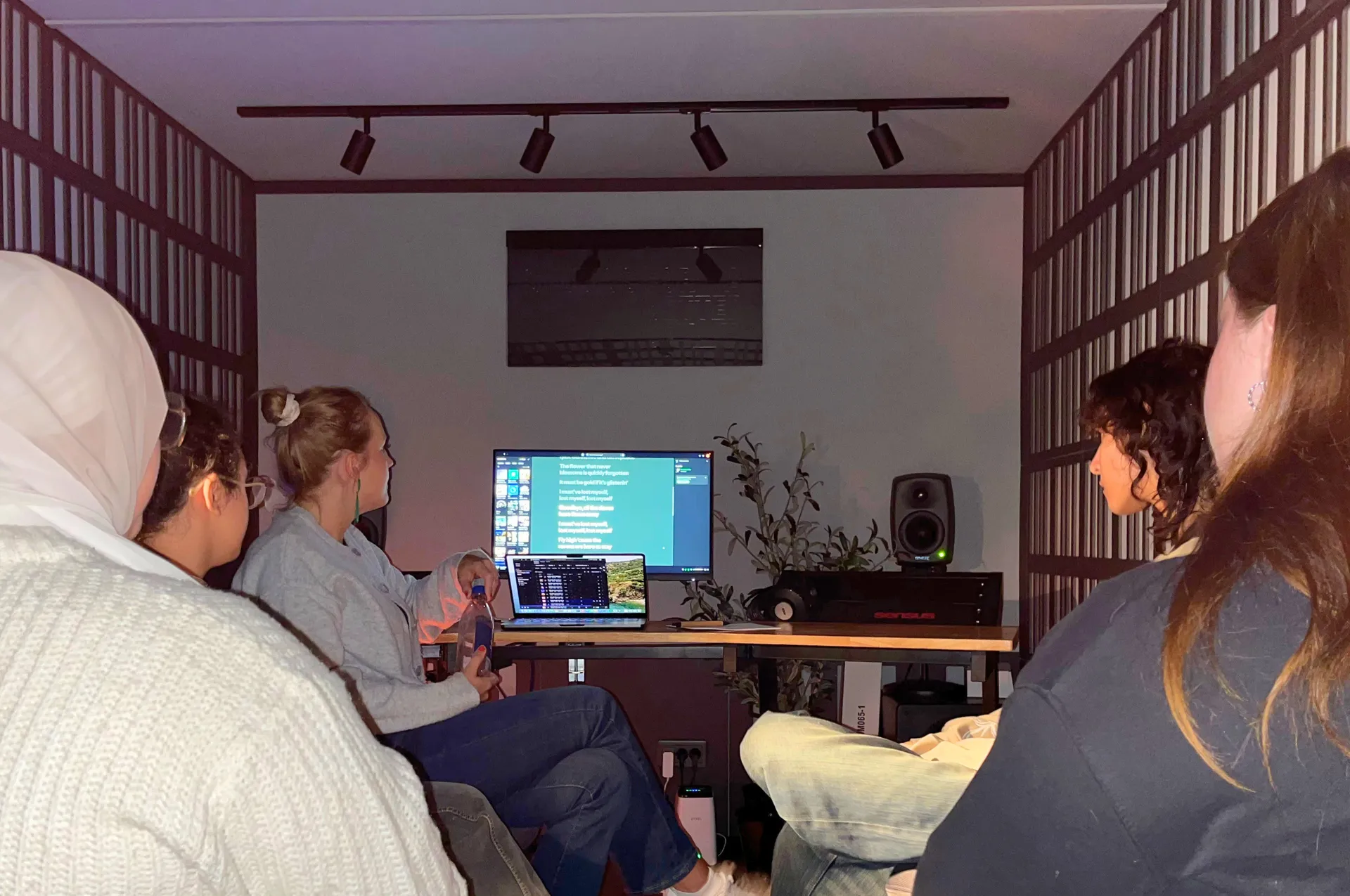 En grupp personer sitter i ett mörkt rum och tittar på en datorskärm med en presentation, i vad som ser ut som en studio eller arbetsplats med högtalare och utrustning