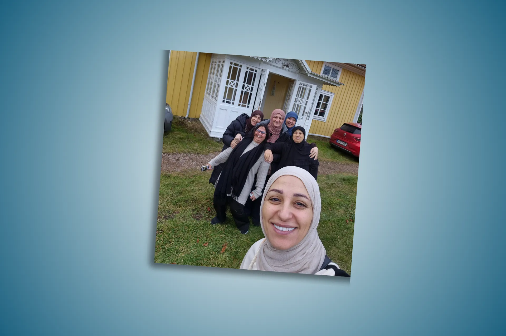 Gruppbild med sex leende kvinnor i hijab som tar en selfie framför ett gult trähus med vita fönster på en grön gräsmatta