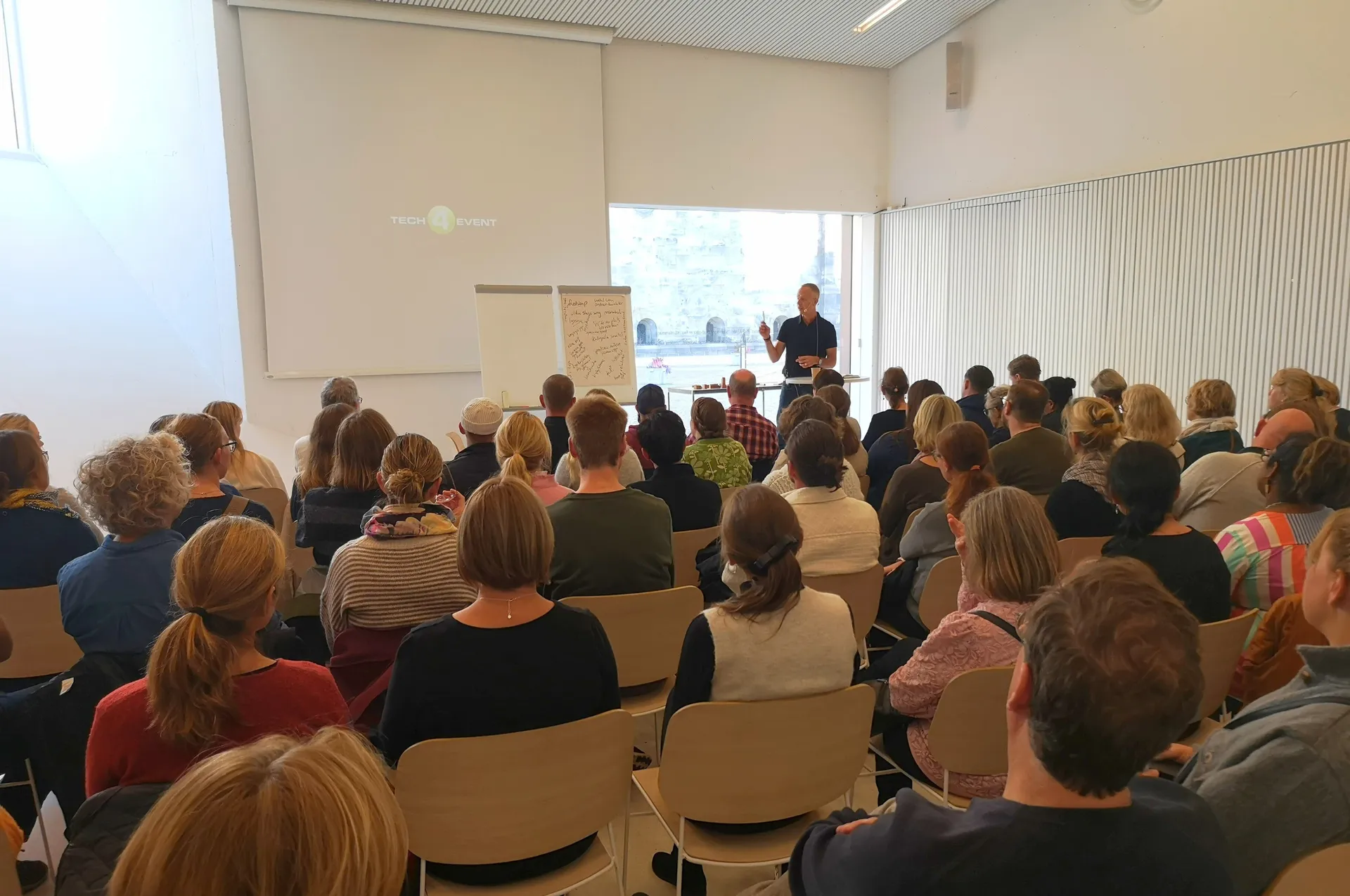 En föreläsare står framför en publik i ett ljust konferensrum och håller en presentation, med whiteboard och blädderblock i bakgrunden