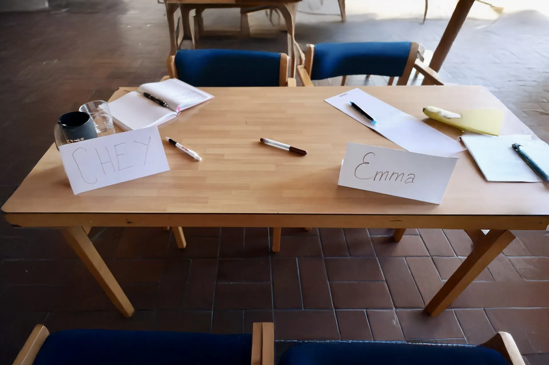 Ett träbord med namnlappar märkta 'Chey' och 'Emma', pennor, ett vattenglas, en kopp och anteckningsblock, förberett för ett möte eller workshop