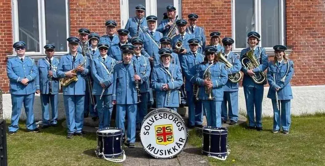 Sölvesborgs Musikkår poserar för gruppfoto utomhus framför en röd tegelbyggnad, klädda i ljusblå uniformer med mössor och hållandes sina blåsinstrument, med en stor bastrumma i förgrunden