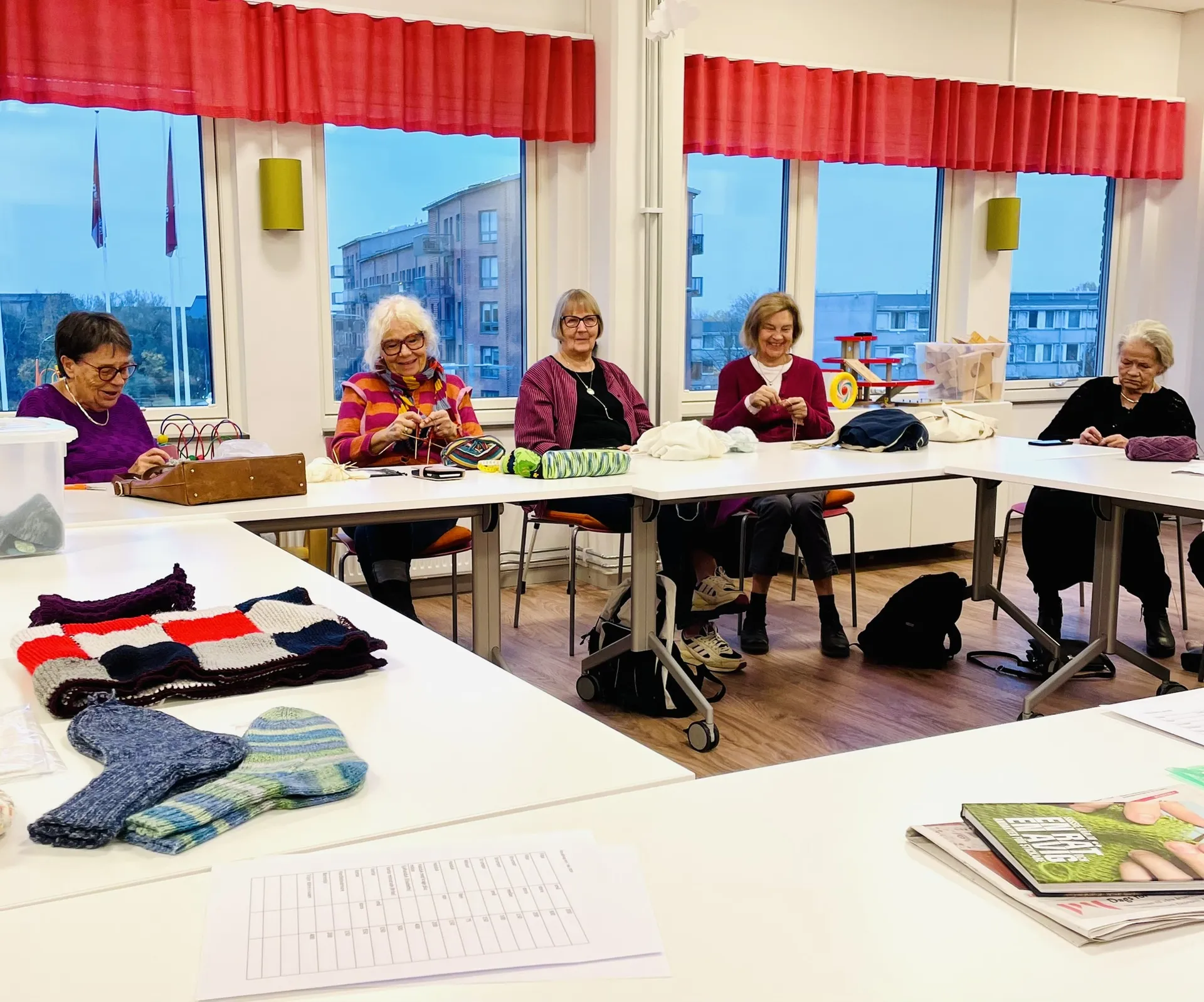 Fem äldre kvinnor sitter vid bord i ett ljust rum och stickar tillsammans. På bordet i förgrunden ligger färdiga stickade alster som vantar och en rutig filt. Röda gardiner ramar in fönstren med utsikt över byggnader.