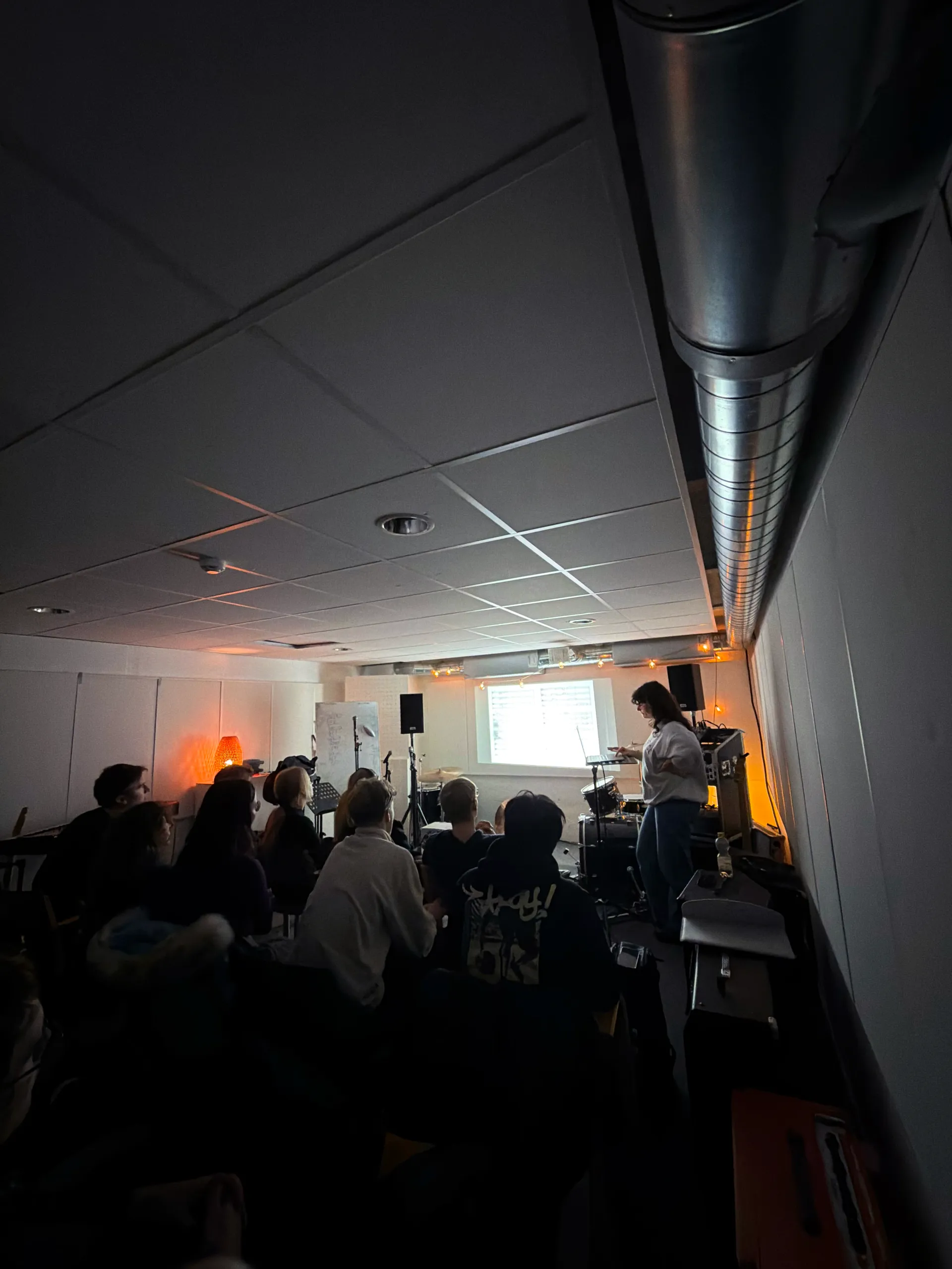 En person håller en presentation framför en grupp åhörare i ett mörkt rum med projektor och varm belysning från en orange lampa
