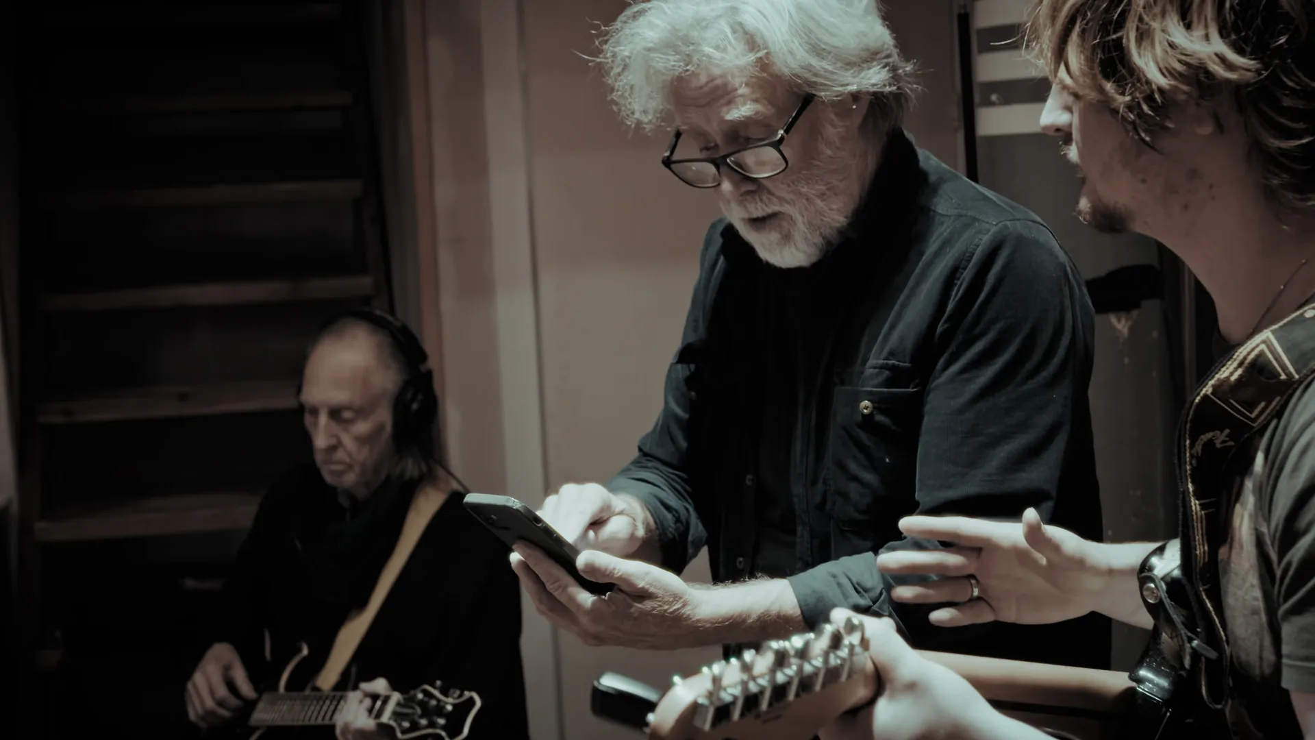 Musiker i en inspelningsstudio där en äldre man med vitt hår och glasögon tittar på en mobiltelefon, medan en annan person håller en gitarr och en tredje person med hörlurar syns i bakgrunden