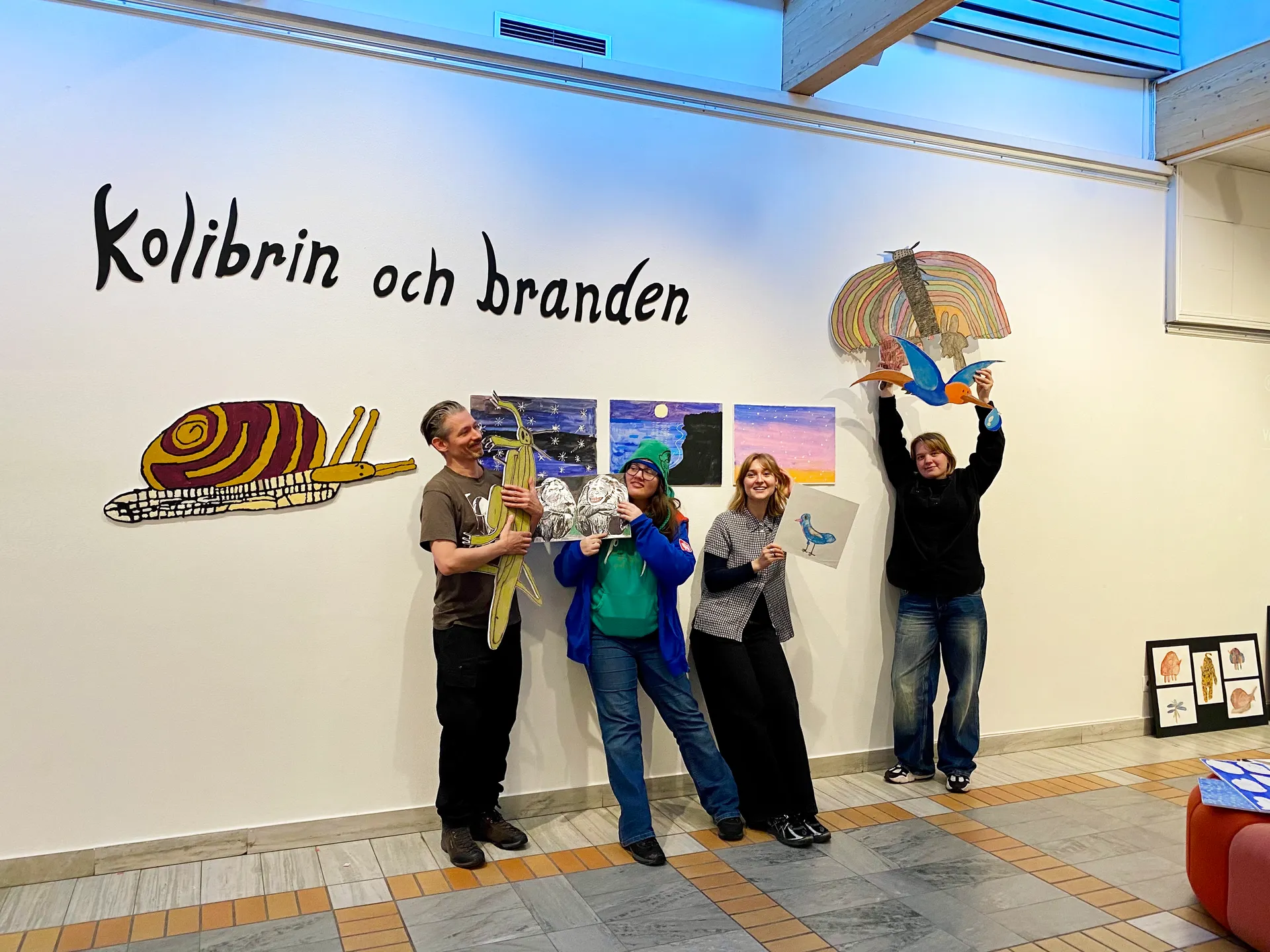 Fyra personer står framför en vit vägg med texten 'Kolibrin och branden' och håller upp färgglada konstverk föreställande djur och natur, inklusive en snigel, gräshoppa, fågel och regnbåge.