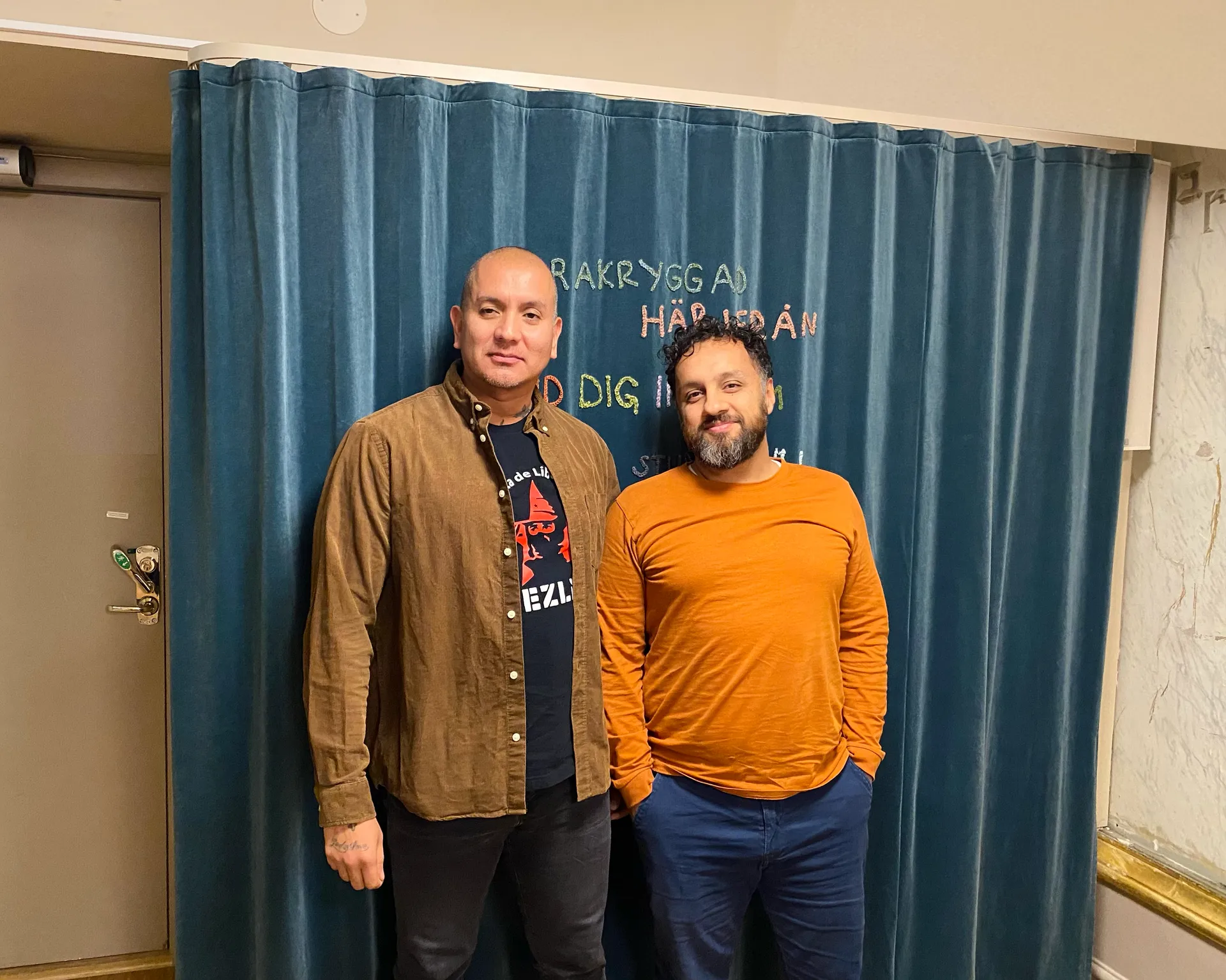 Två män står framför ett blått draperi med text. Mannen till vänster har rakad skalle och bär en brun skjorta över en mörk t-shirt. Mannen till höger har mörkt lockigt hår och skägg och bär en orange tröja.