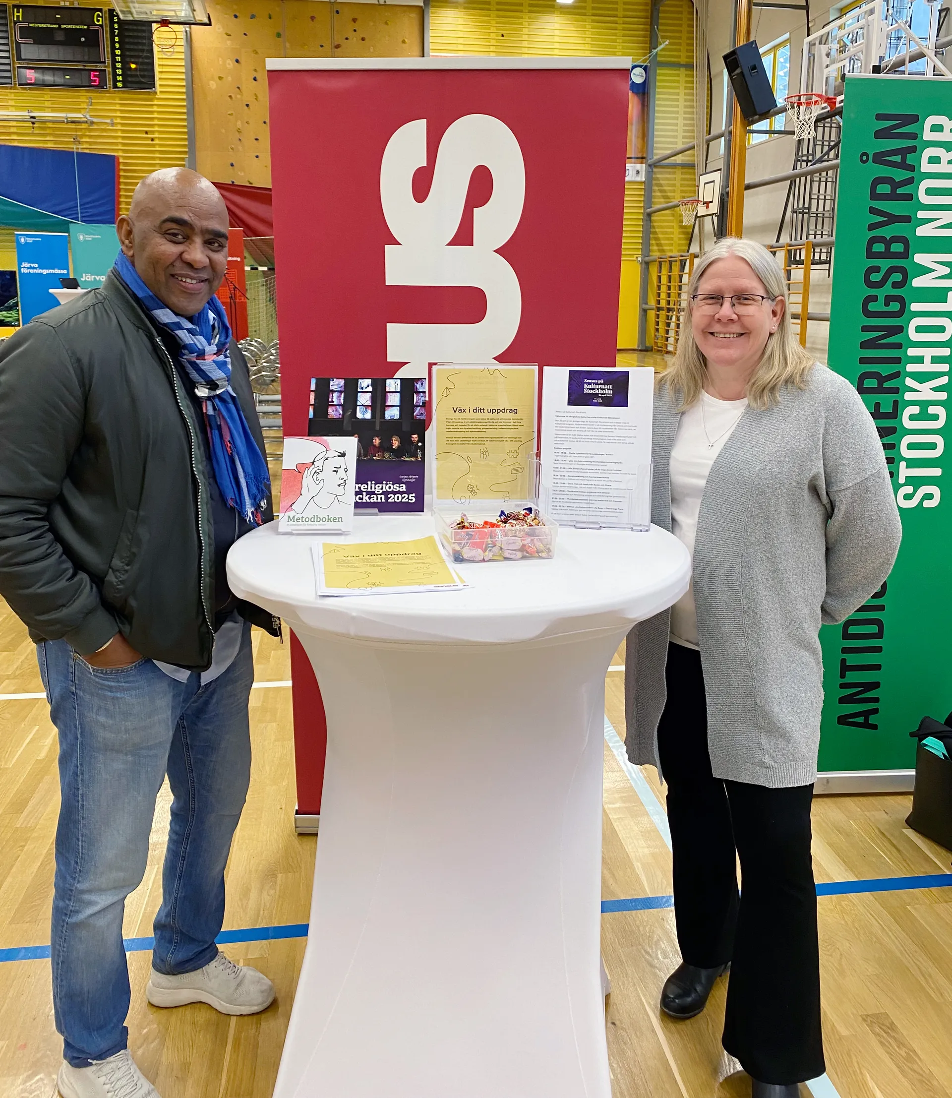 Två personer står vid ett informationsbord i en idrottshall. På bordet finns broschyrer om Interreligiösa veckan 2025 och annat material. Bakom dem syns en röd roll-up med Sensus logotyp och en grön roll-up för Föreningsbyrån Stockholm Norr.
