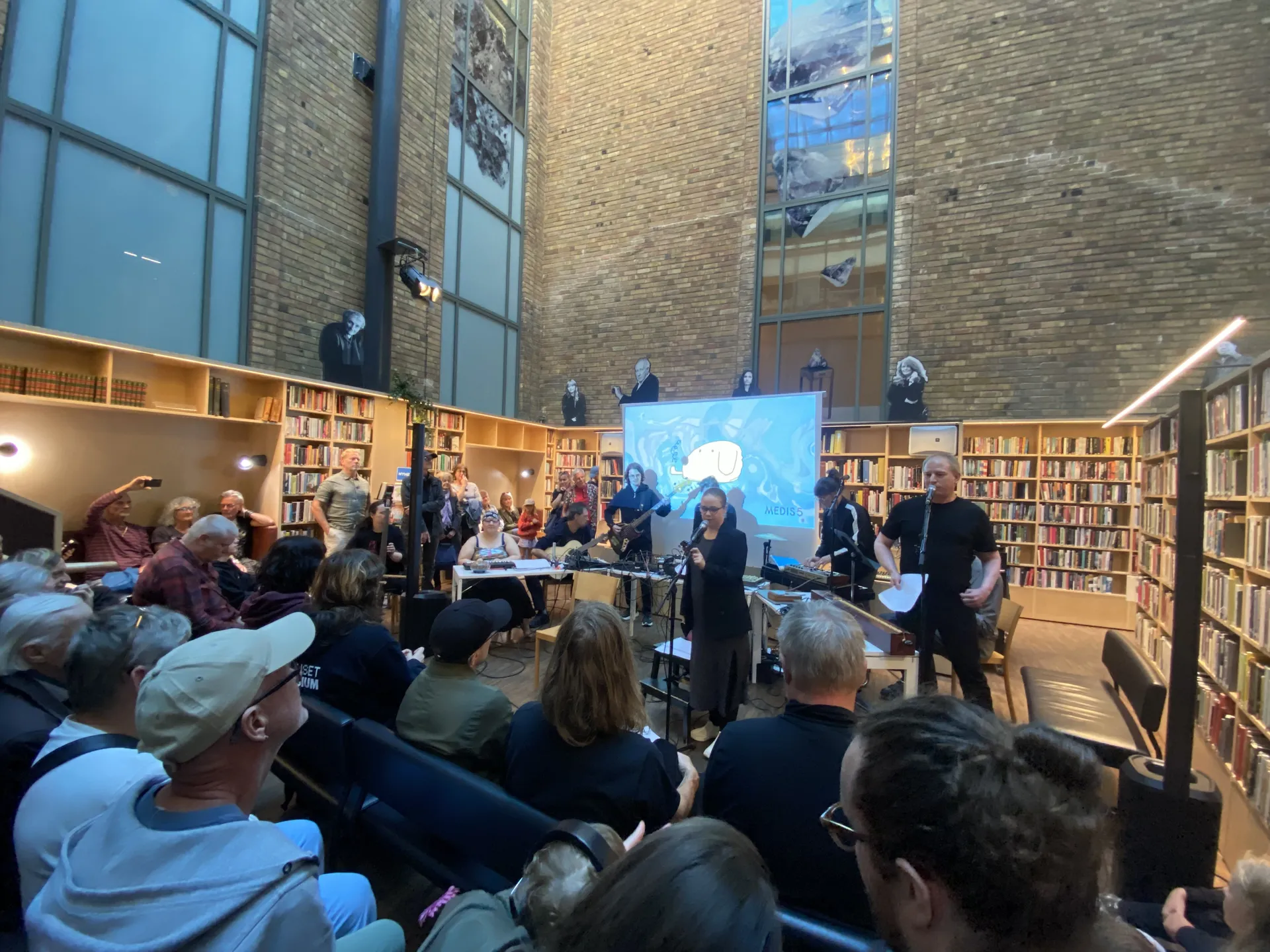 Musikframträdande i ett bibliotek med höga tegelväggar och bokhyllor, där musiker uppträder framför en sittande publik med en projicerad bild i bakgrunden