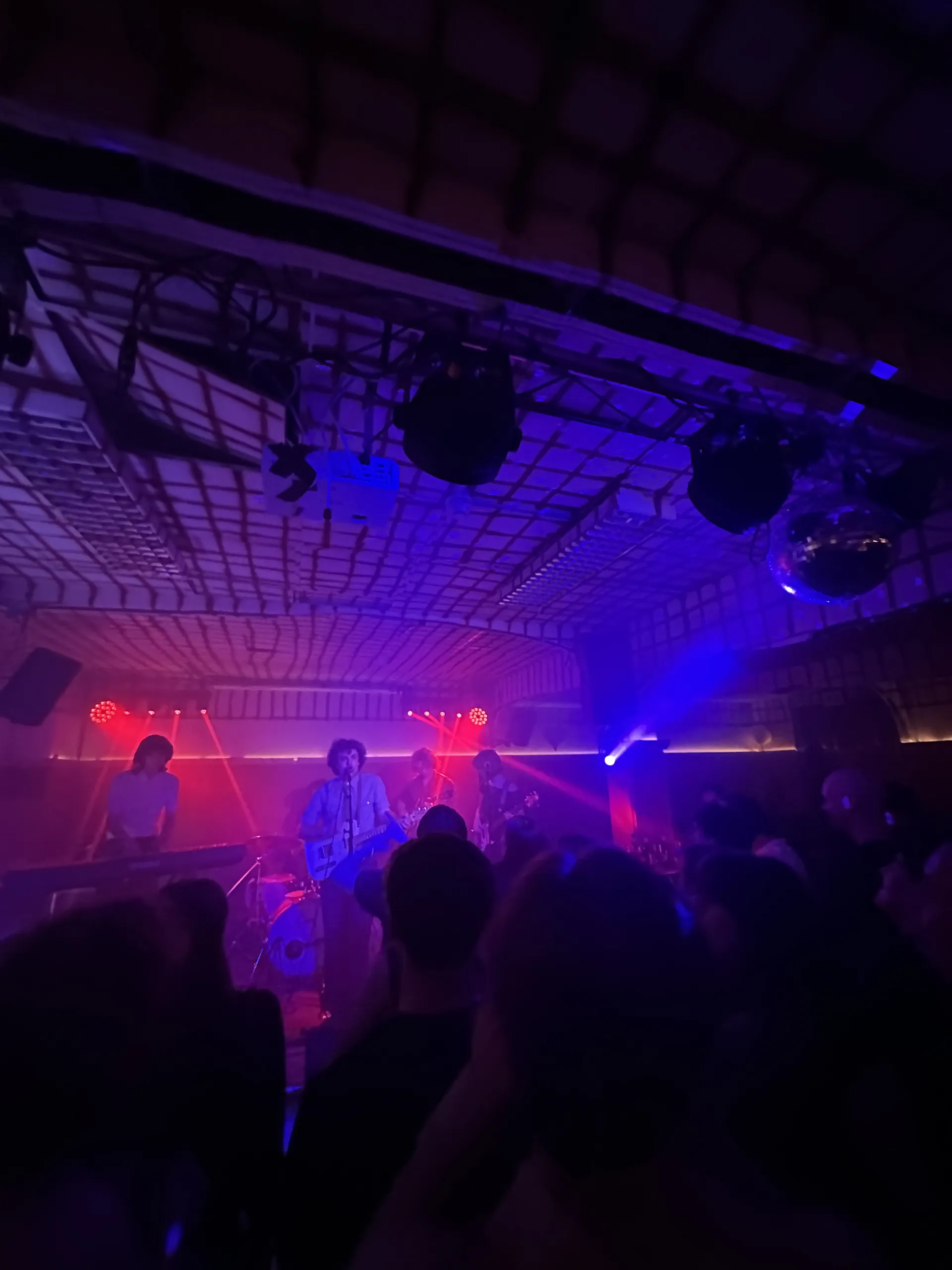 Ett liveband spelar på en scen i en mörk konsertlokal med blå och röd scenbelysning, medan publiken står och tittar på framträdandet