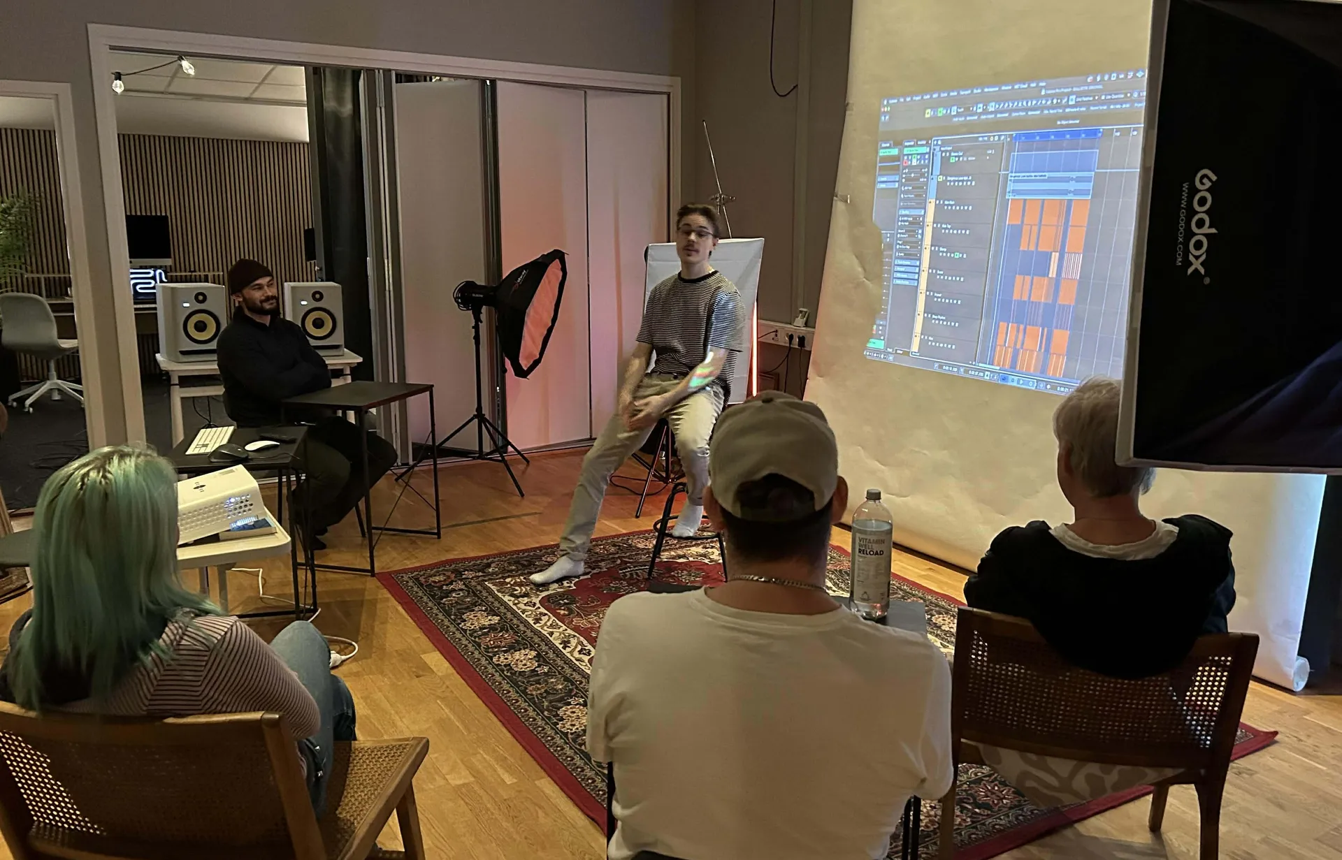 En ung man håller en workshop eller presentation om musikproduktion i en studio, med ett musikprogram projicerat på väggen medan flera deltagare sitter och lyssnar