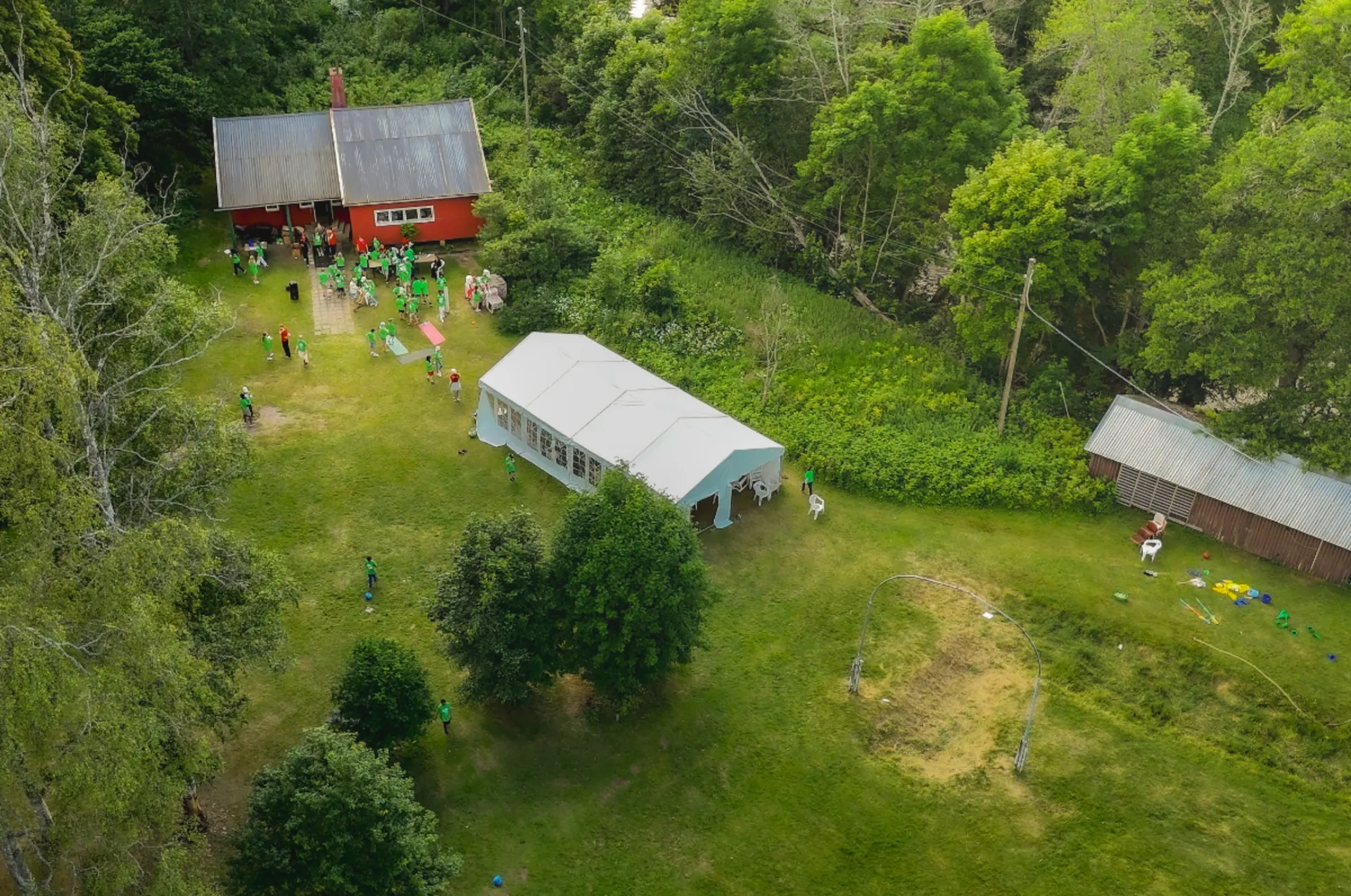 Flygbild över en lägerplats med röd stuga, vitt partytält och människor i gröna tröjor som samlas på en gräsmatta omgiven av lummig skog