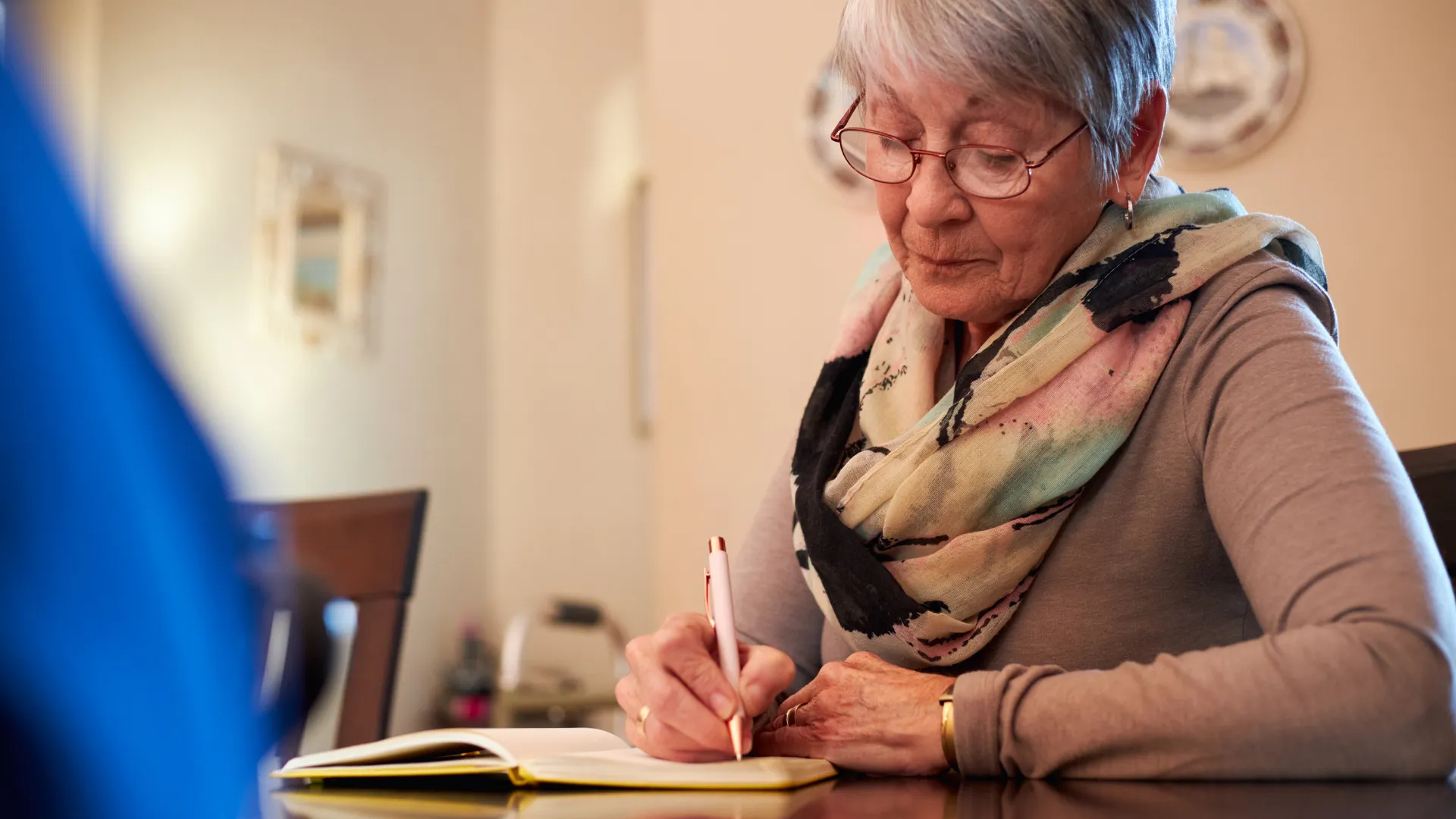 Äldre kvinna med grått hår och glasögon sitter vid ett bord och skriver i en anteckningsbok med en penna, iklädd beige tröja och färgglad sjal