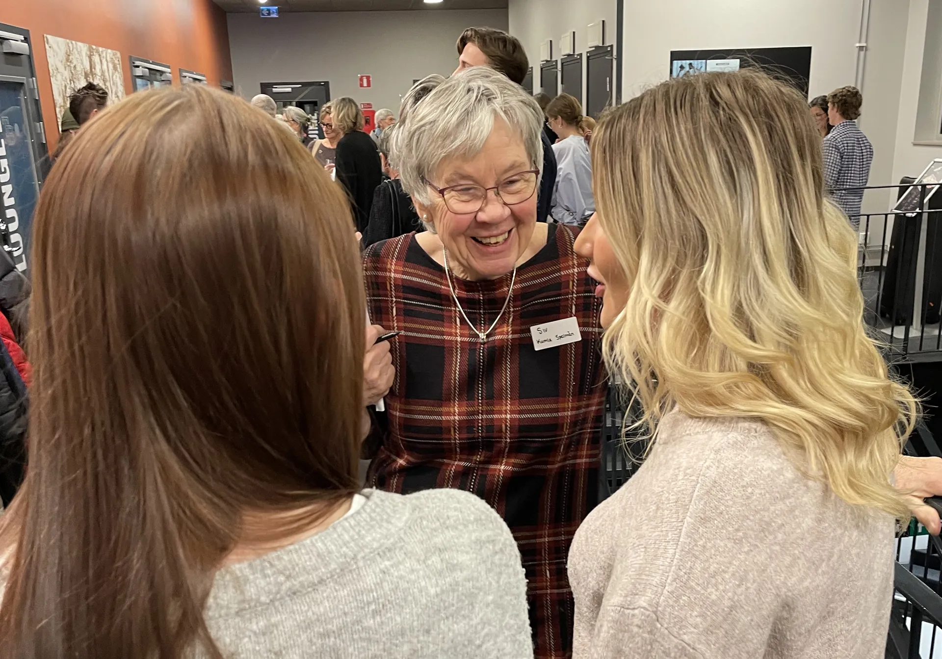 En äldre kvinna med grått hår och glasögon ler och samtalar med två yngre kvinnor på ett socialt evenemang. Hon bär en rutig tröja och en namnskylt. I bakgrunden syns fler personer som minglar.