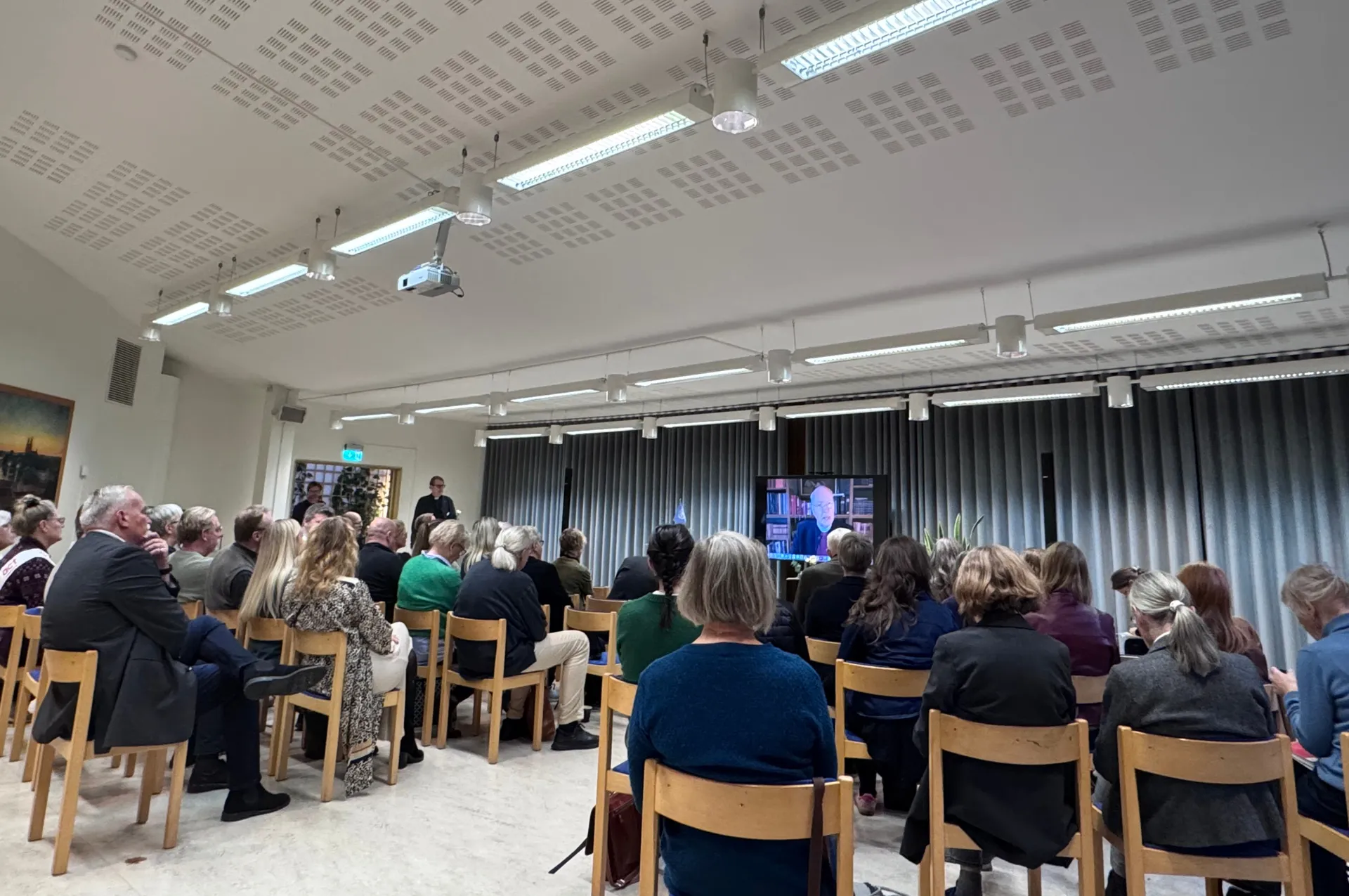 Publik sitter på trästolar i en ljus konferenssal och tittar på en videolänk med en talare som visas på en stor skärm framme vid grå gardiner