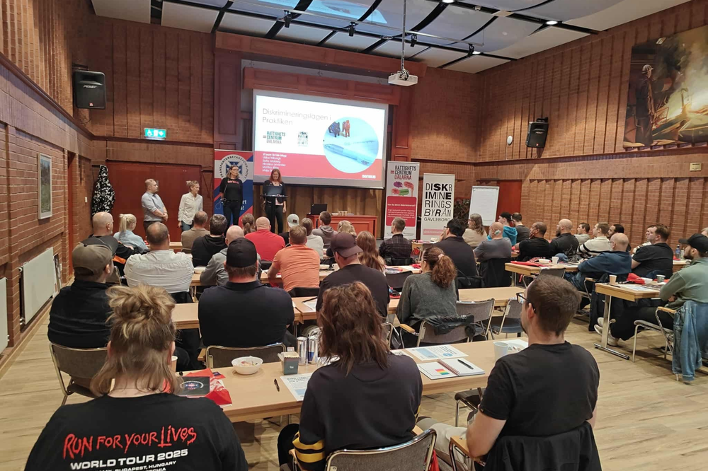 Ett fullsatt mötesrum där flera personer sitter vid bord och lyssnar på en presentation om diskrimineringslagen. Fyra föreläsare står längst fram, och bakom dem visas en projicerad presentation samt roll‑ups från olika organisationer.