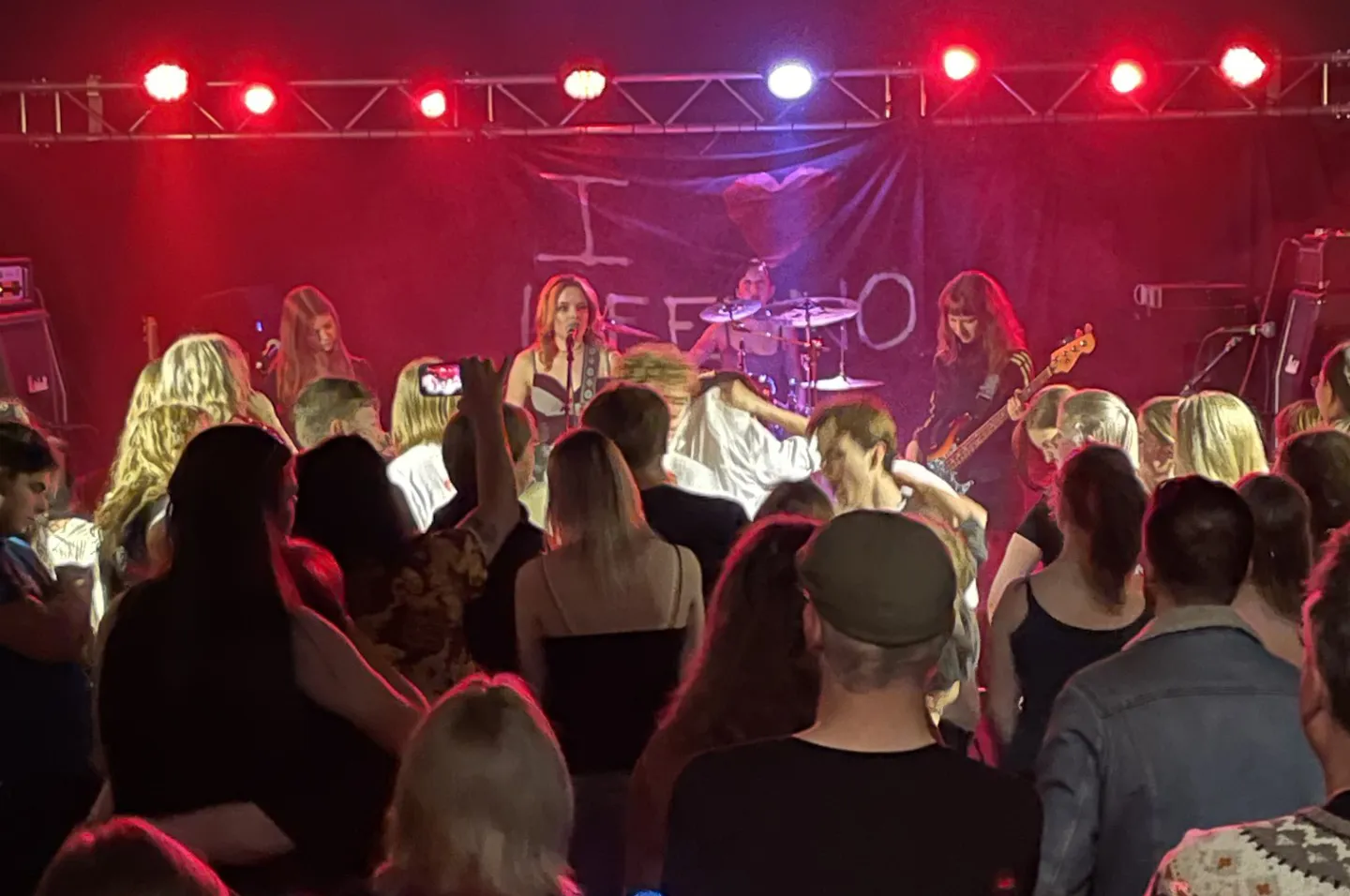 Ett rockband uppträder på en scen med röd belysning framför en publik. Bandet består av flera medlemmar inklusive en sångerska, gitarrist och trummis, med en banderoll i bakgrunden.