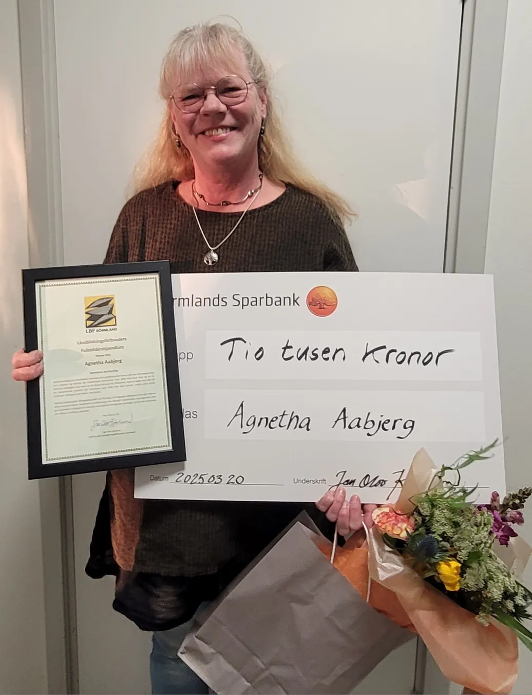 Agnetha Aabjerg håller upp ett inramat diplom från Länsbildningsförbundets Folkbildarstipendium och en stor check på tio tusen kronor från Sörmlands Sparbank, tillsammans med en blombukett.
