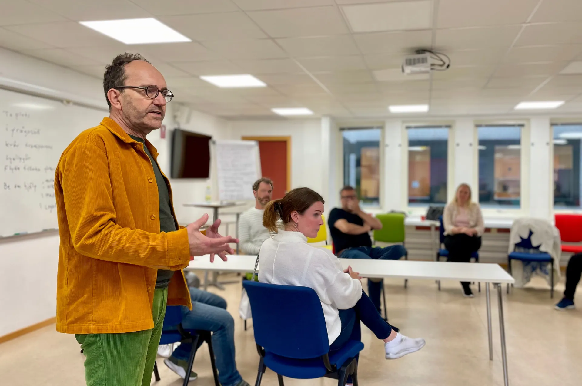 En man i gul manchesterjacka och glasögon håller en presentation eller föreläsning i ett konferensrum medan flera personer sitter och lyssnar på stolar runt om i rummet.