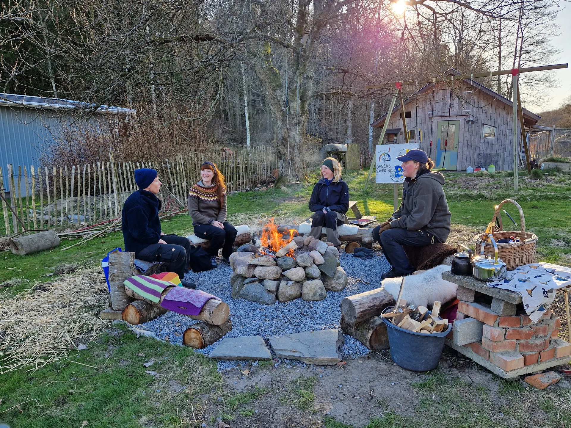 Fyra personer sitter runt en brasa på stockar i en trädgård under tidig vår eller sen höst, med träbyggnader och kala träd i bakgrunden