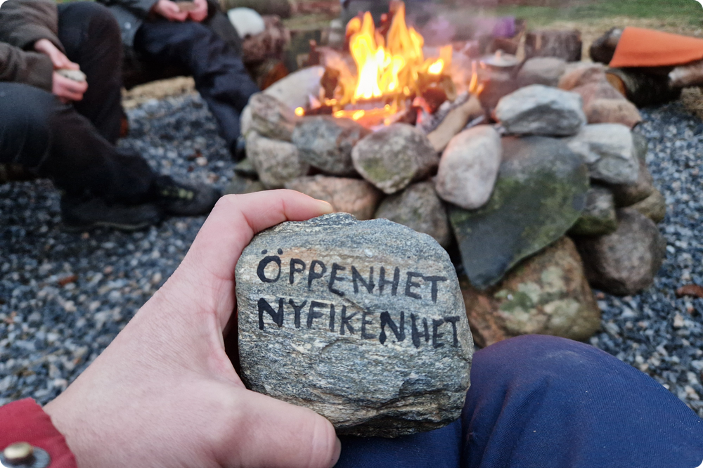 En hand håller upp en sten där orden "ÖPPENHET NYFIKENHET" står inristade. I bakgrunden gräs och man anar en eldstad.