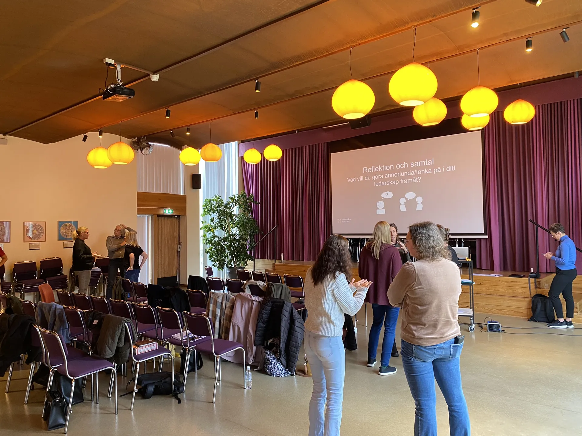 Konferenslokal med deltagare som minglar under en paus. En projektorduk visar texten 'Reflektion och samtal' om ledarskap. Rummet har lila gardiner, orangea taklampor och rader av stolar.