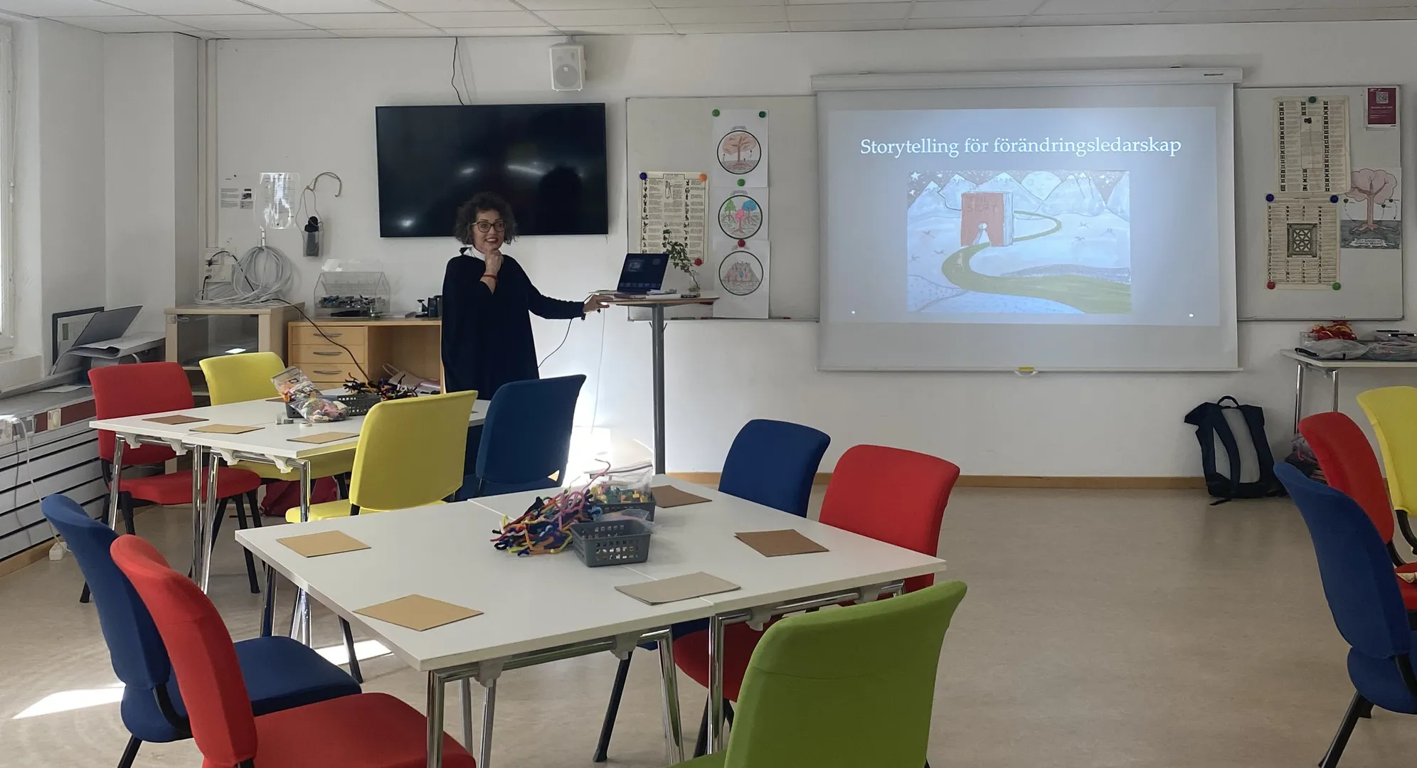 En kvinna håller en presentation om storytelling för förändringsledarskap i ett klassrum med färgglada stolar i rött, blått, gult och grönt. På projektorduken visas en illustration med texten 'Storytelling för förändringsledarskap'.