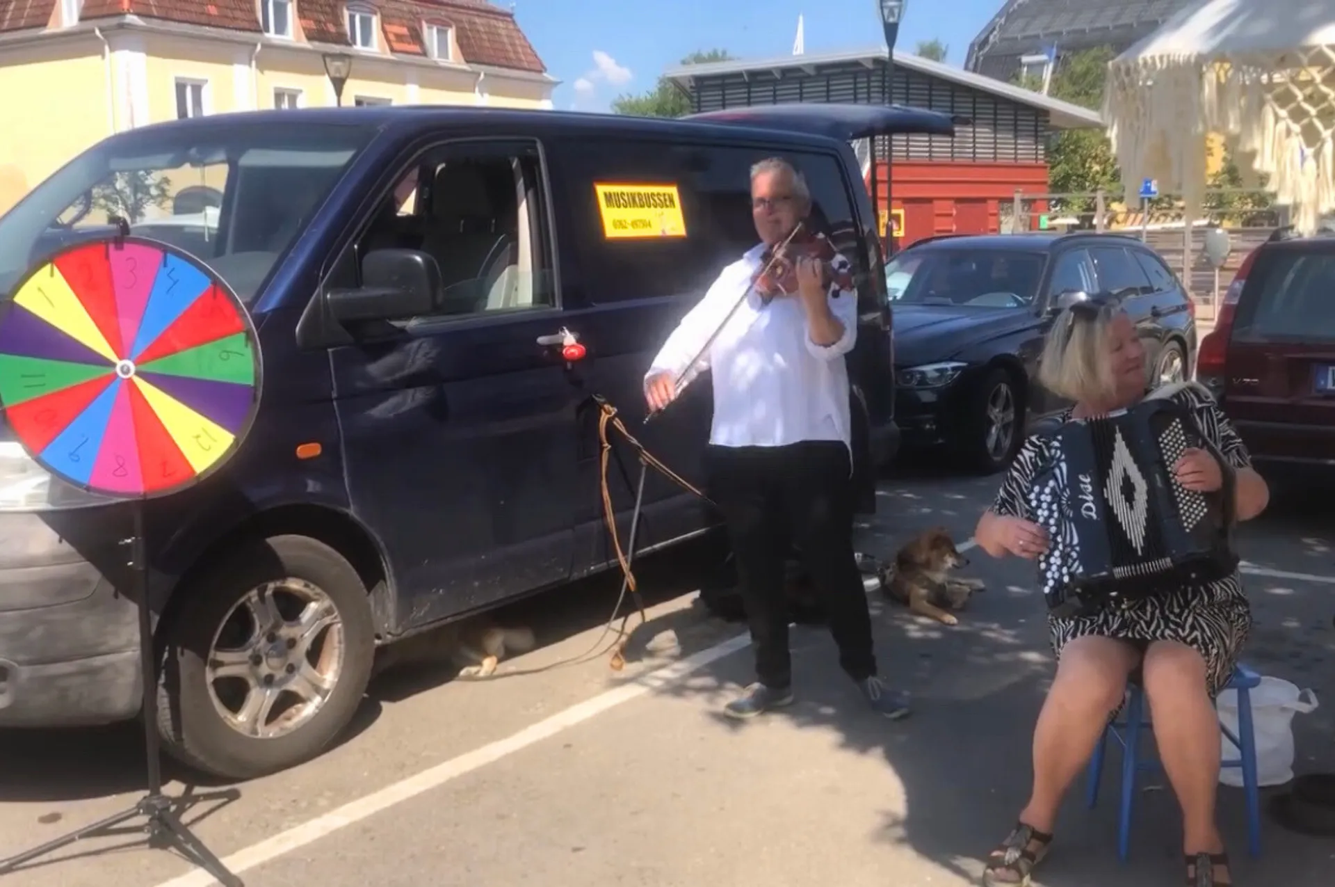Två gatumusikanter spelar musik utomhus vid en mörk minibuss märkt Musikbussen. En man spelar fiol och en kvinna spelar dragspel. Ett färgglatt lyckohjul står bredvid bussen och en hund ligger på marken.