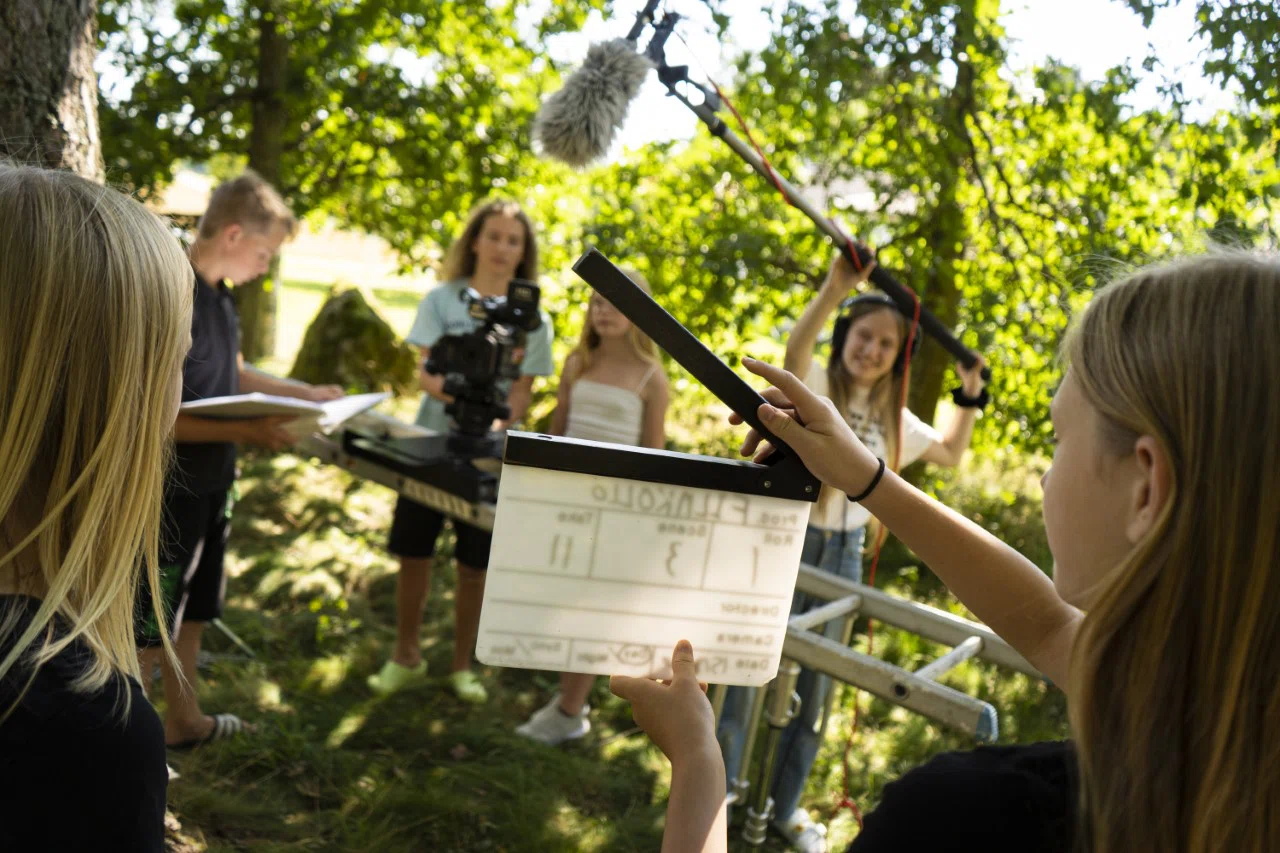 Ungdomar spelar in en film utomhus i grön natur. En person håller en filmklappa i förgrunden medan andra hanterar kamera och bommikrofon i bakgrunden.