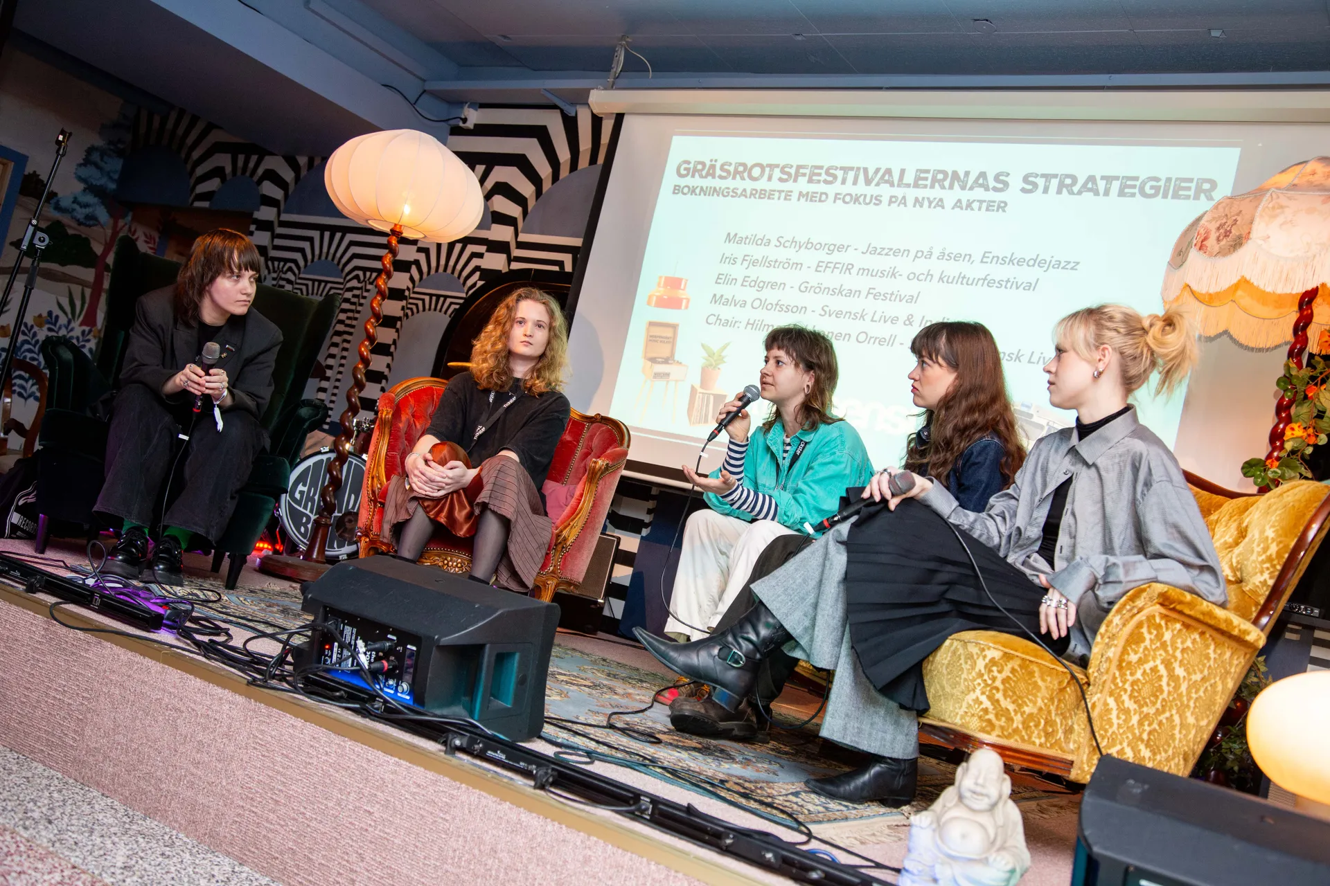 Fem kvinnor sitter i en paneldiskussion om gräsrotsfestivalernas strategier för bokningsarbete med fokus på nya akter. De sitter i bekväma fåtöljer och soffor på en scen med en presentation projicerad bakom dem.