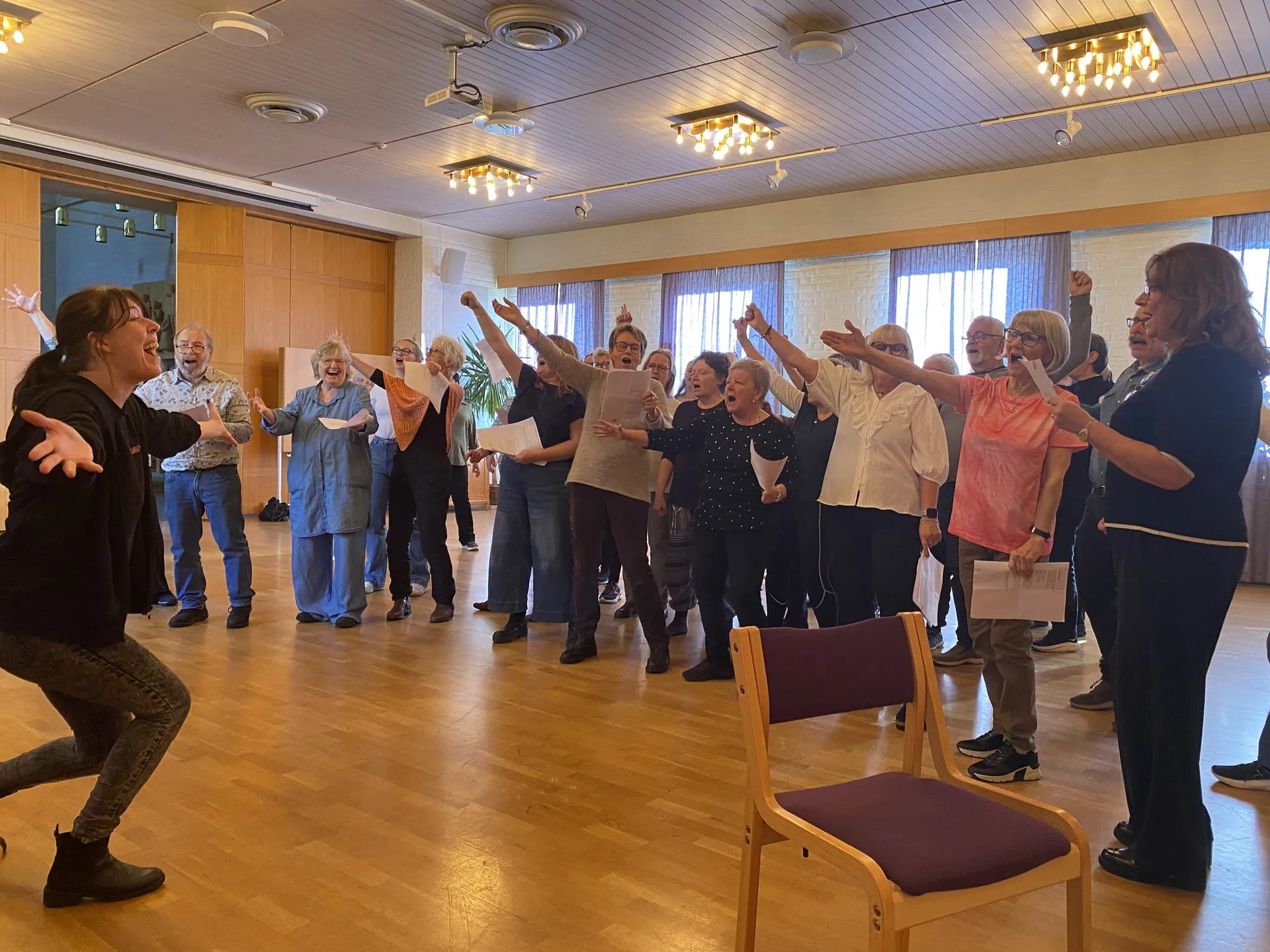 En kör med vuxna personer övar tillsammans i en ljus samlingssal. Deltagarna håller notblad och sträcker entusiastiskt upp armarna medan en körledare till vänster dirigerar gruppen.