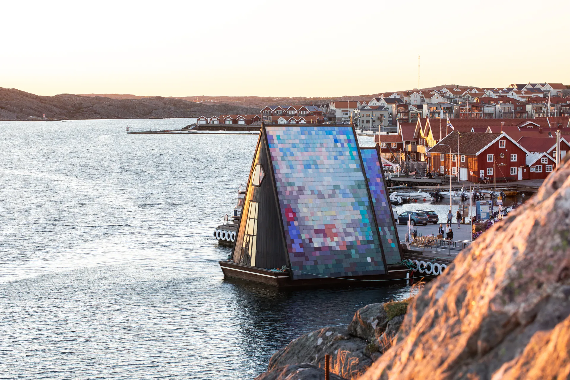 Flytande arkitektonisk struktur med färgglad mosaikfasad i en svensk skärgårdshamn med traditionella röda sjöbodar i bakgrunden vid solnedgång
