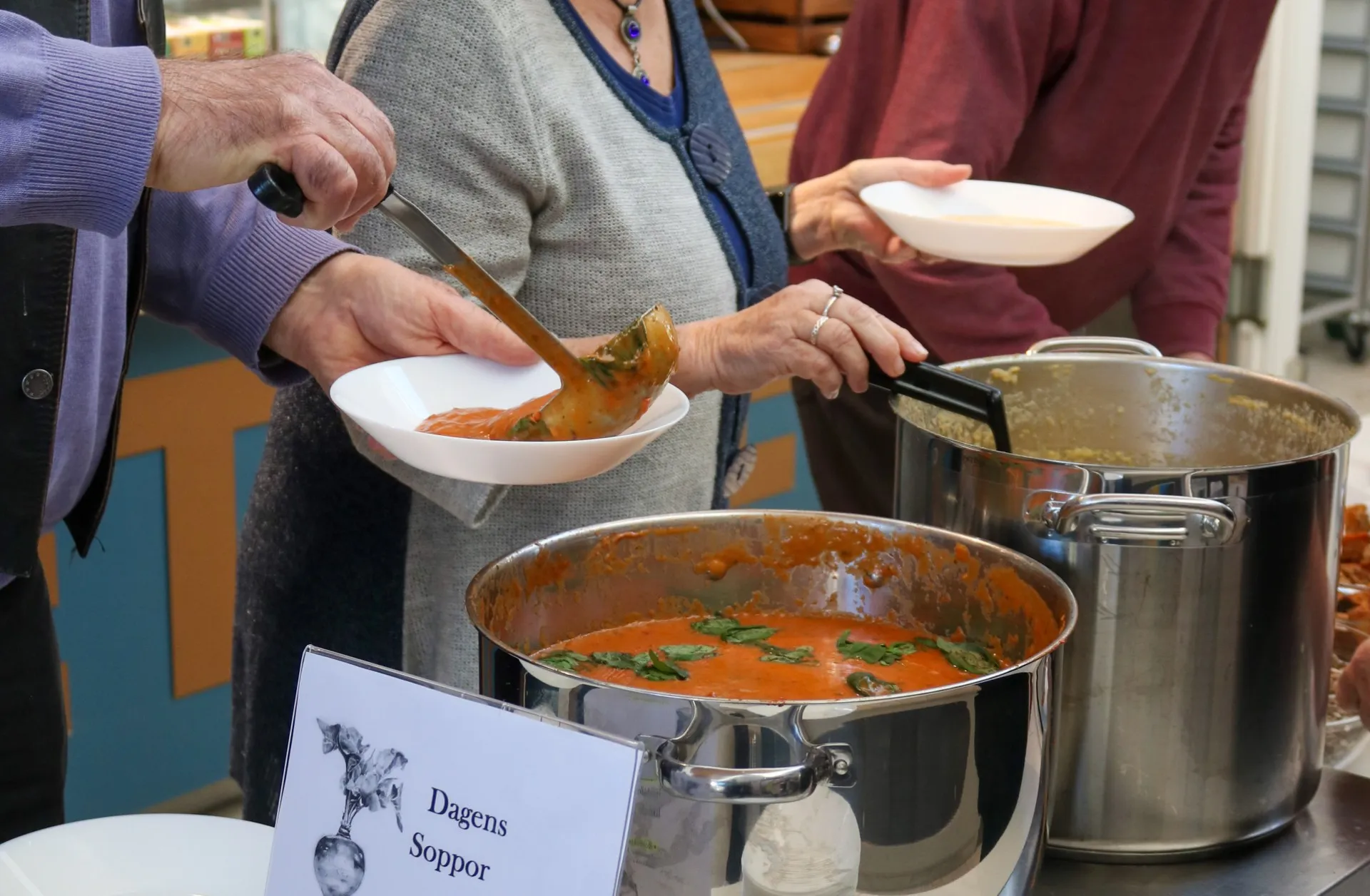 Personer serverar sig själva soppa från stora grytor vid en sopplunch. En skylt med texten 'Dagens Soppor' syns framför grytorna med tomatsoppa garnerad med basilika.