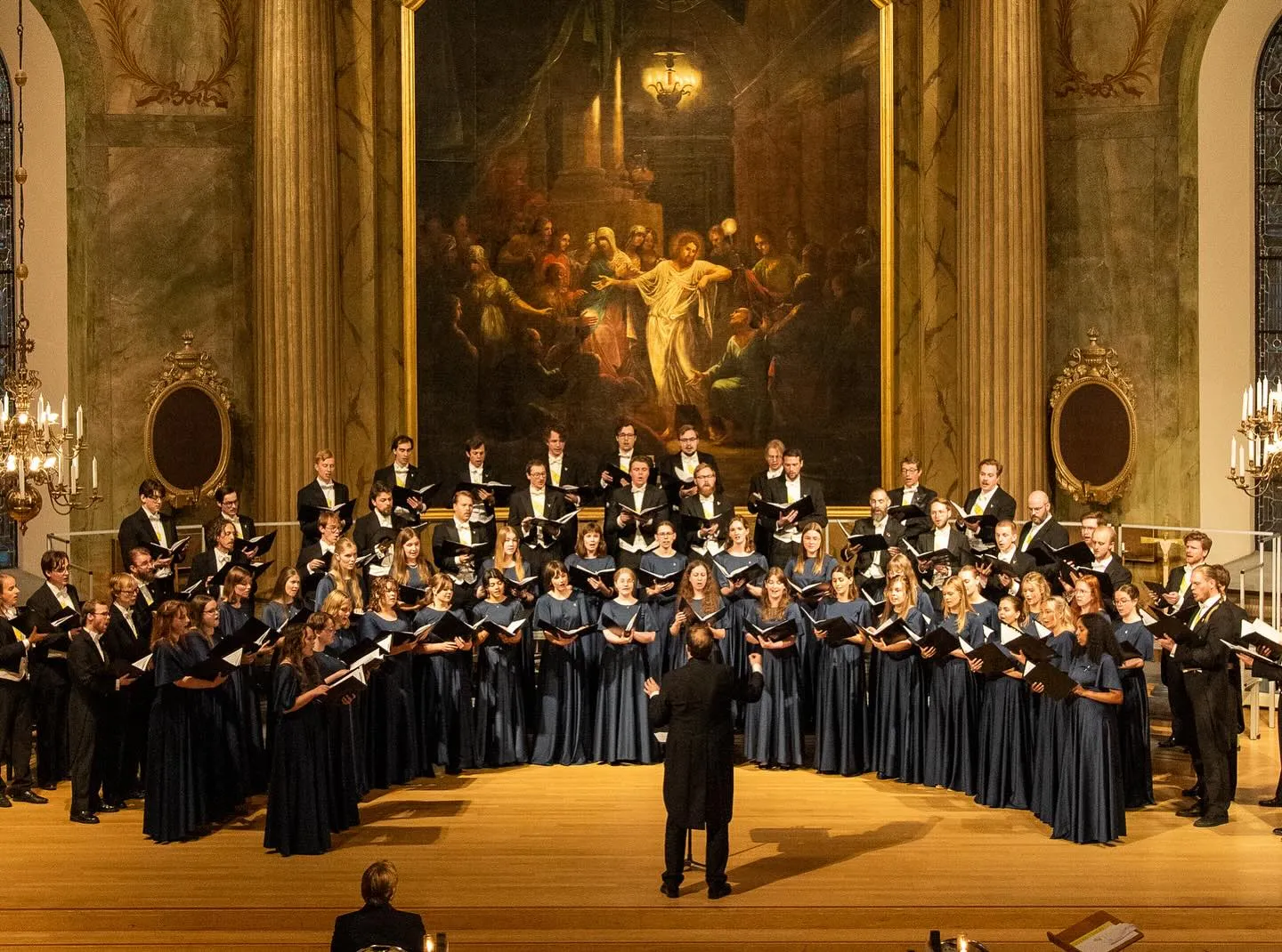 En stor kör uppträder i en prålig kyrka med en stor religiös målning i bakgrunden. Kvinnorna bär mörkblå klänningar och männen svarta kostymer med gula detaljer. En dirigent leder kören från mitten av scenen.