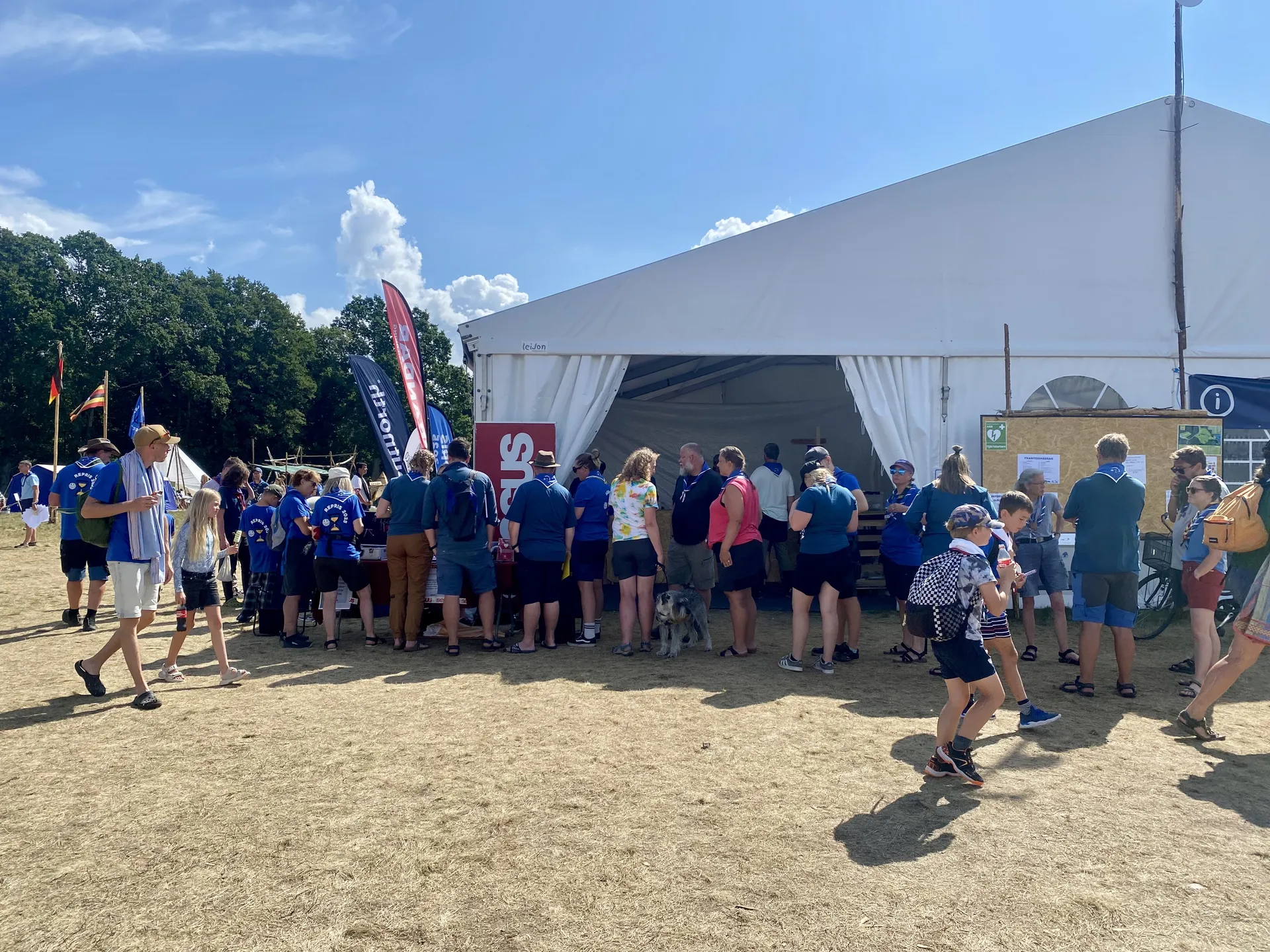 Människor i scoutuniformer köar vid ett stort vitt tält på ett soligt scoutläger, med flaggor och informationsskyltar synliga i bakgrunden