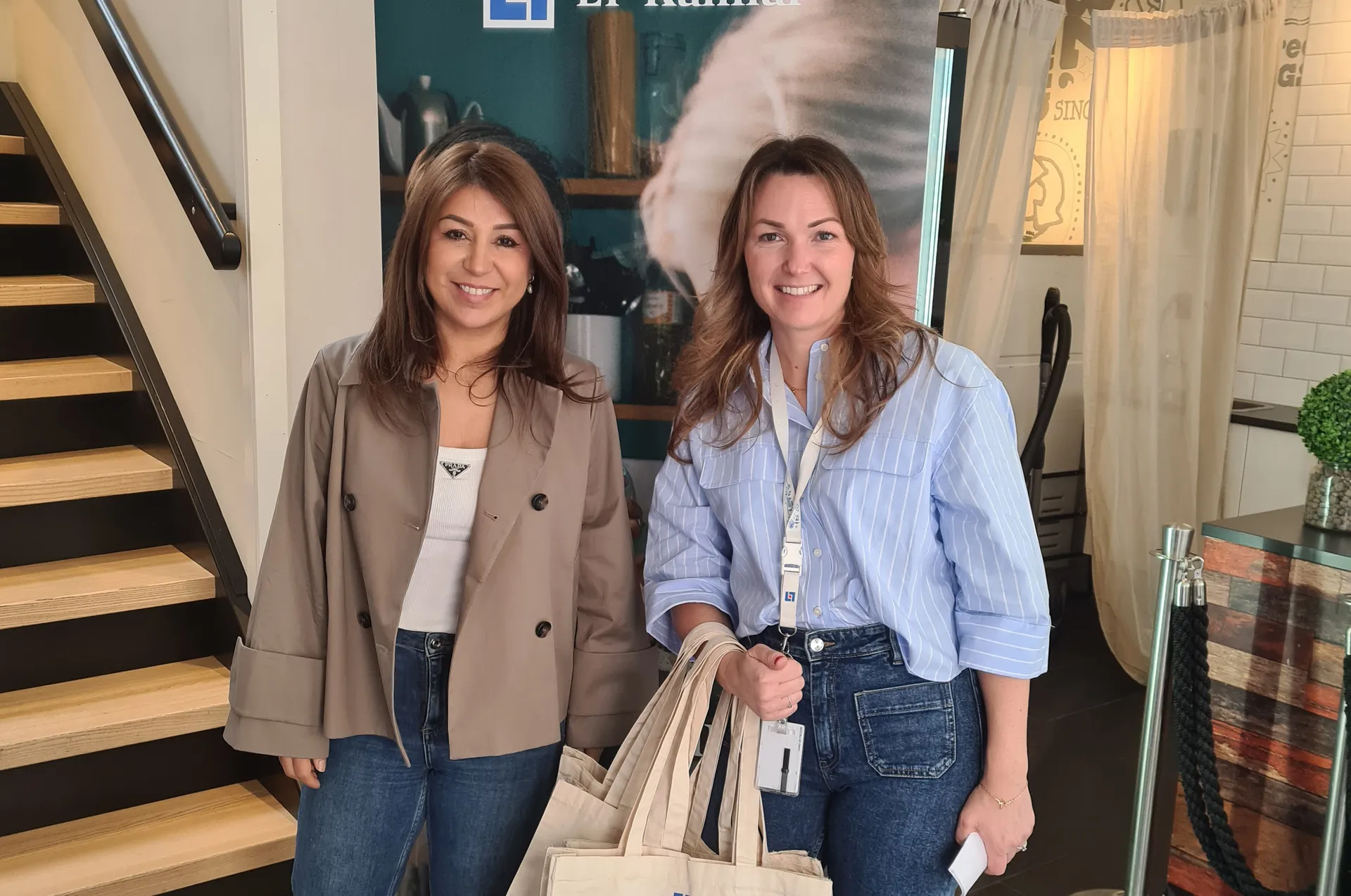 Två leende kvinnor står tillsammans vid en mässmonter. Den ena bär en beige kavaj och den andra en ljusblå randig skjorta och håller i tygkassar. I bakgrunden syns en trappa och en reklamaffisch.