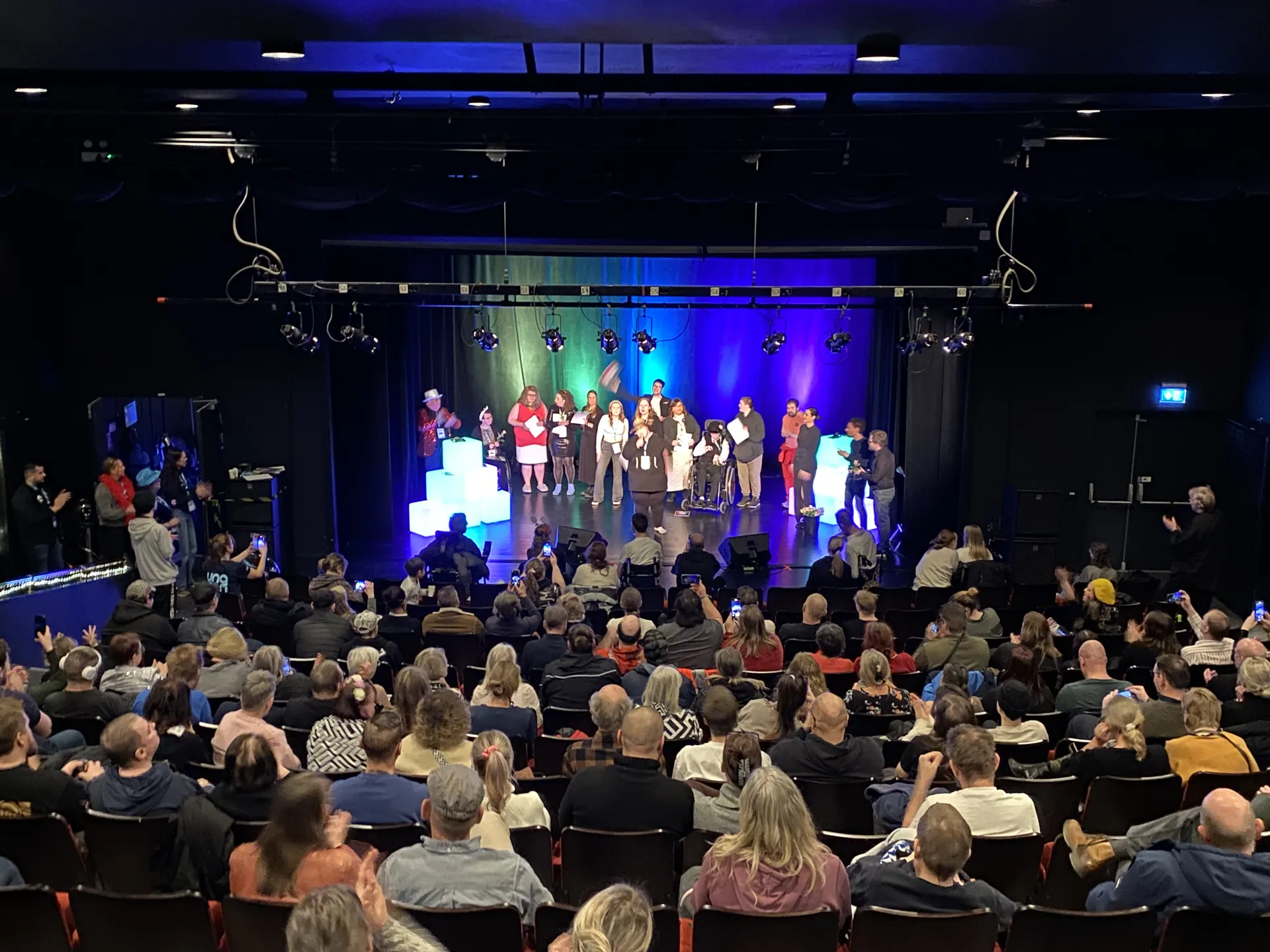 En grupp människor står på en upplyst scen framför en sittande publik i en mörk teaterlokal, med blå och grön scenbelysning i bakgrunden