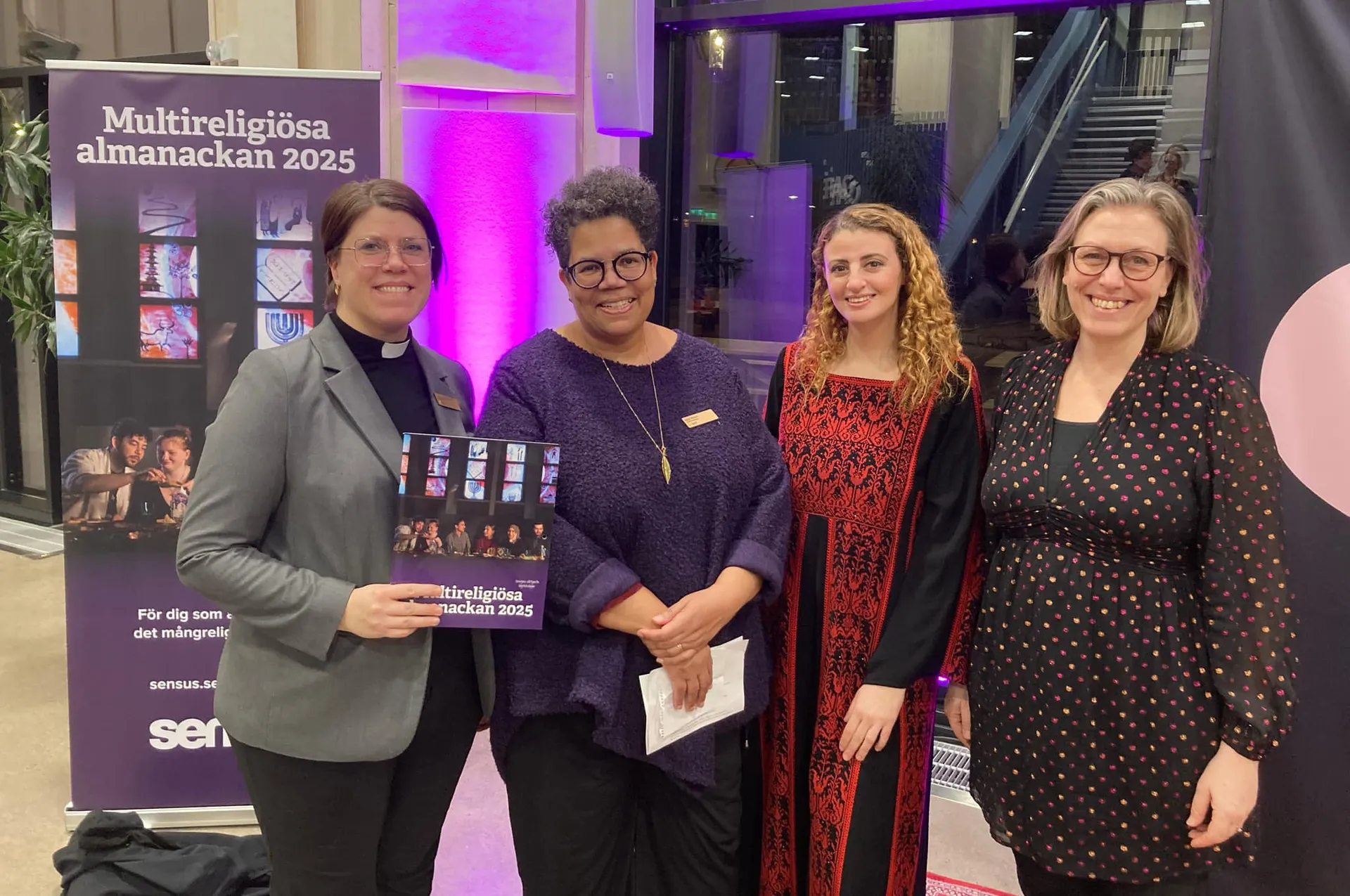 Fyra leende kvinnor poserar tillsammans vid lanseringen av Multireligiösa almanackan 2025. En av kvinnorna håller i almanackan och bär prästkrage. I bakgrunden syns en roll-up med information om almanackan och lila belysning.
