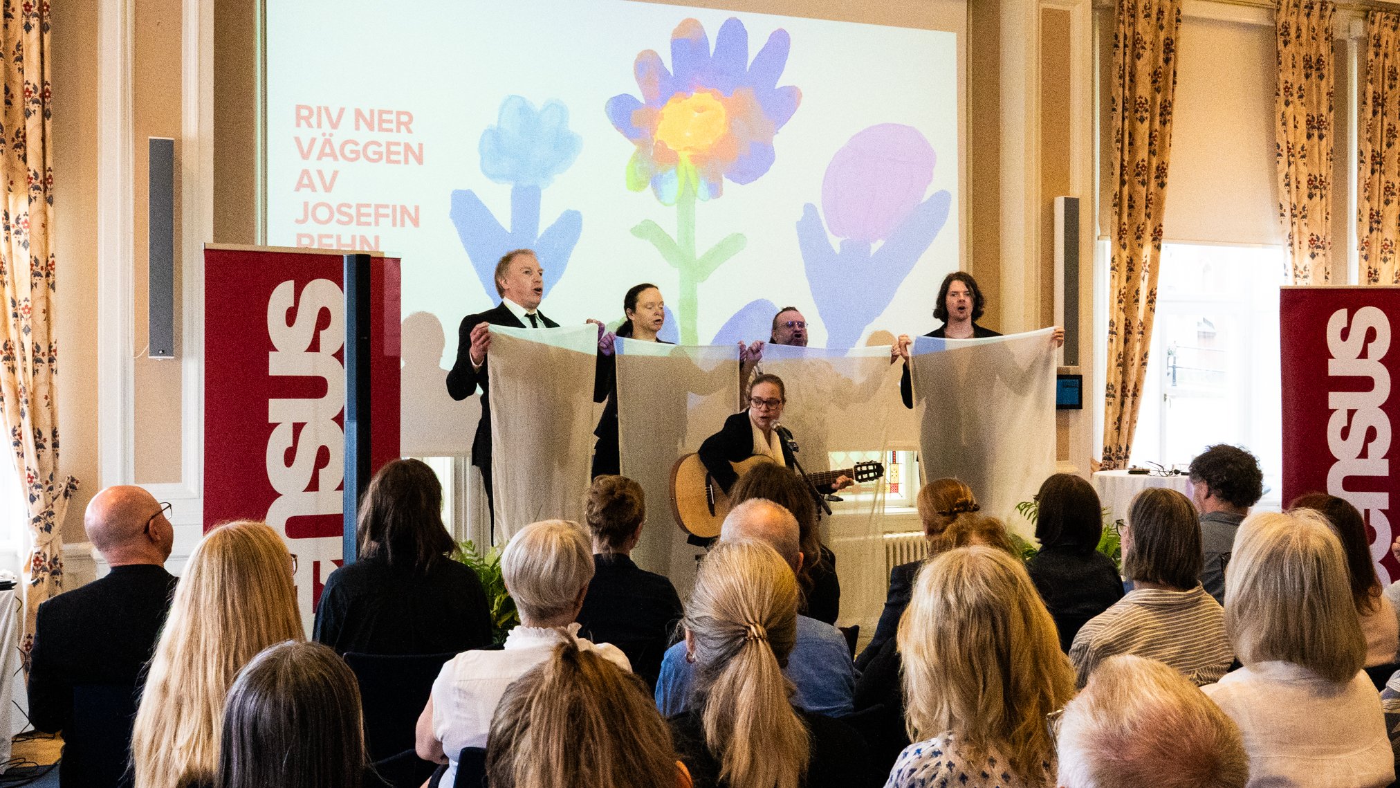 Fyra personer står längst fram i en sal och håller upp ett tygstycke medan de sjunger. En person sitter framför och spelar gitarr. På väggen projiceras texten ”Riv ner väggen” och färgglada blommor. Publik sitter vänd mot scenen.