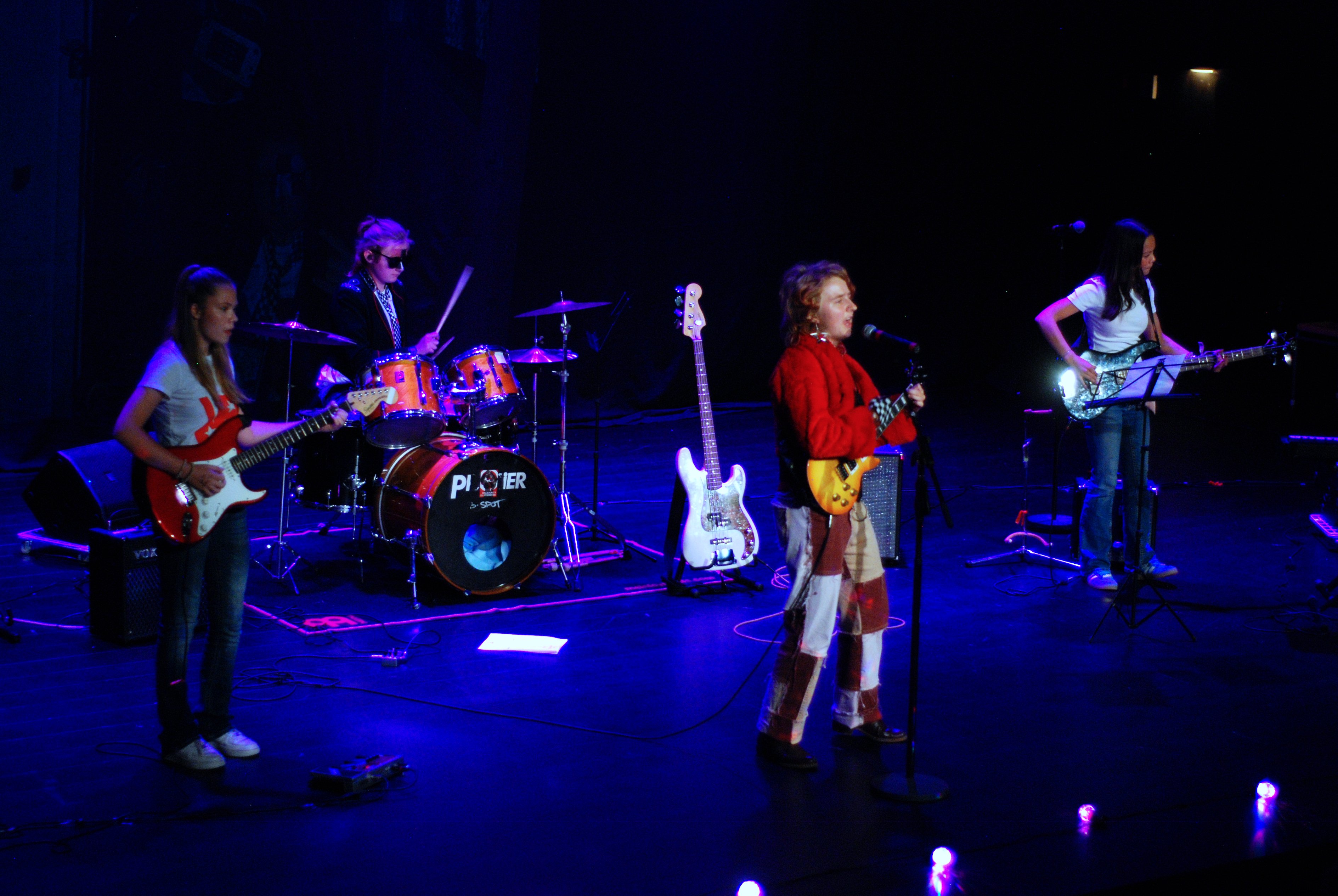 Fyra unga musiker uppträder på en scen med blå och lila scenljus. De spelar elgitarrer, bas och trummor, och en av dem sjunger i mikrofon. Musikutrustning och instrument syns runtom på scenen.