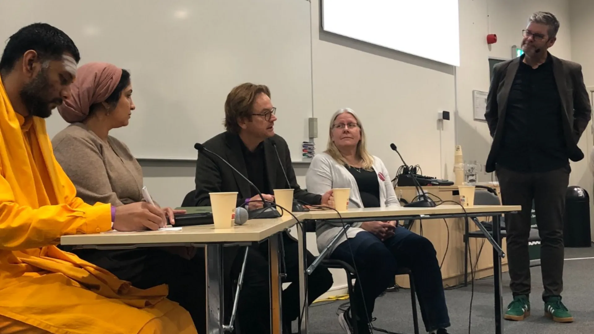 Paneldiskussion i ett konferensrum med fem personer sittande vid ett bord med mikrofoner och kaffemuggar, medan en man i mörk kavaj står och talar till höger