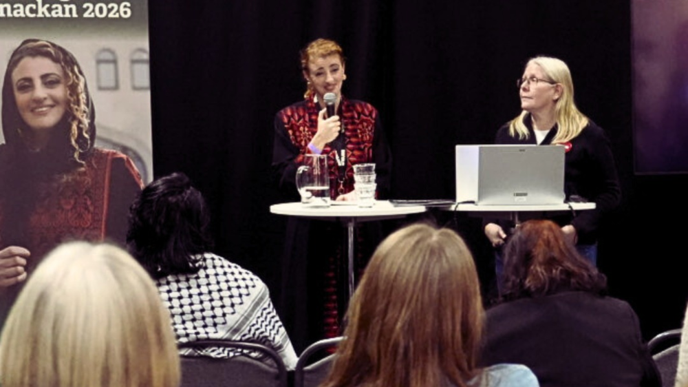 Två personer står på scen vid ett runt bord under ett seminarium. Den ena talar i mikrofon, den andra sitter vid en laptop. I förgrunden syns publik. Bredvid scenen står en affisch med porträtt och texten ”Almanackan 2026”.