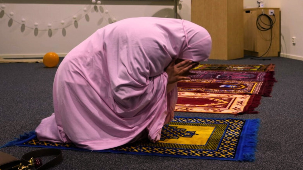 En person i ljuslila klädsel knäböjer i bön på en färgglad bönematta i ett stilla rum. Flera bönemattor ligger utlagda på golvet, vilket ger intryck av en gemensam plats för stillhet och reflektion.