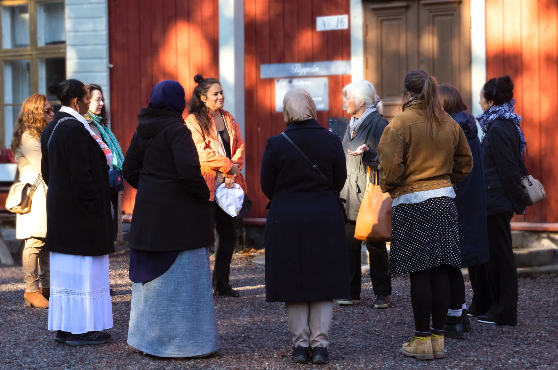 En grupp kvinnor i olika åldrar och med olika kulturella bakgrunder står och samtalar utomhus framför en röd träbyggnad med vita fönsterkarmar, troligen på en kulturhistorisk plats i Sverige.