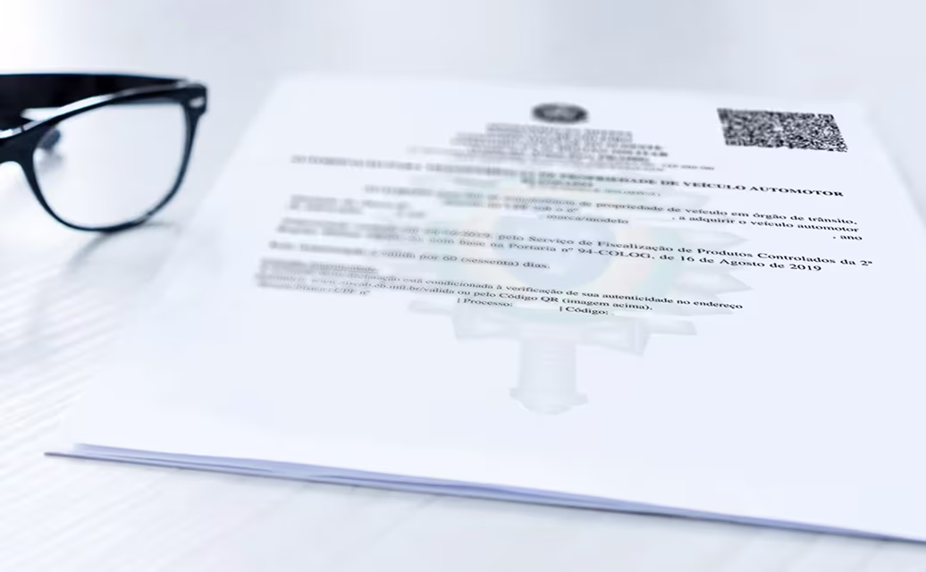 Documento oficial de veículo automotor em papel branco ao lado de um óculos preto sobre uma mesa branca.