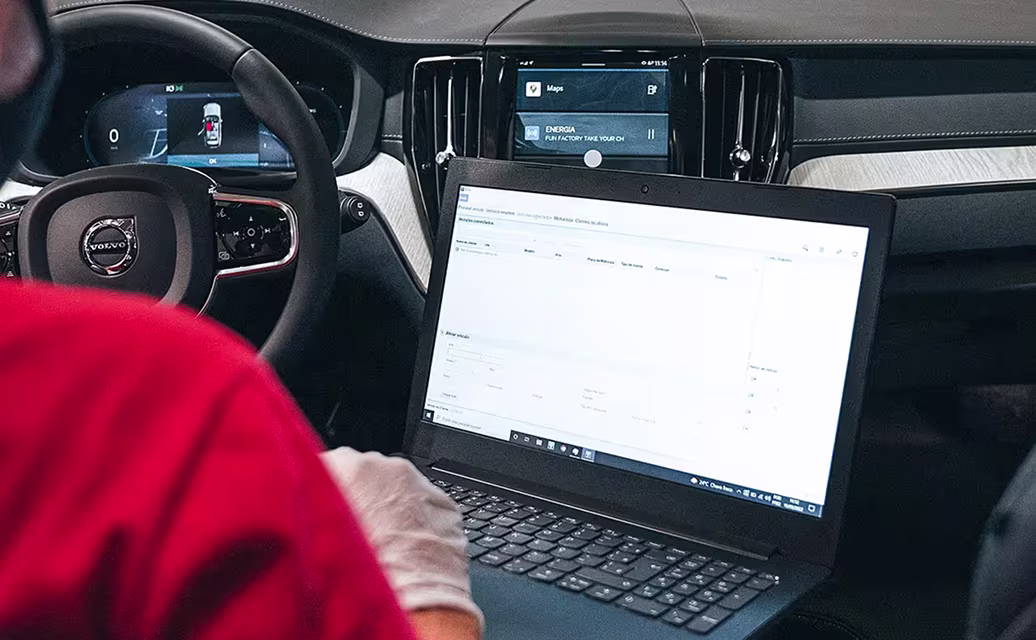 Pessoa com luva branca usando laptop dentro de um carro Volvo, mostrando o volante e a tela do sistema de navegação do veículo.